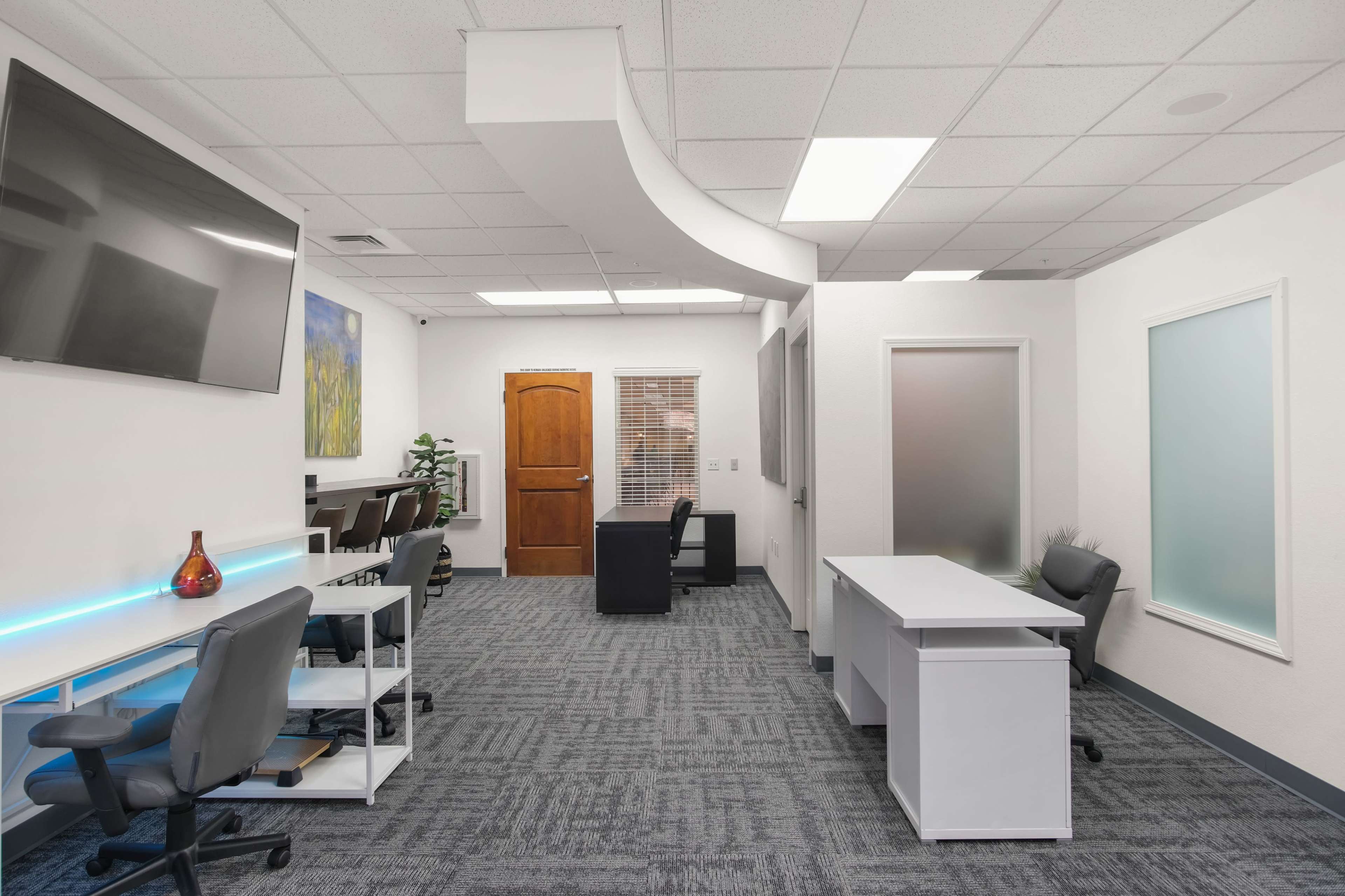 A modern office space with multiple workstations, a wooden door, and a large television mounted on the wall.