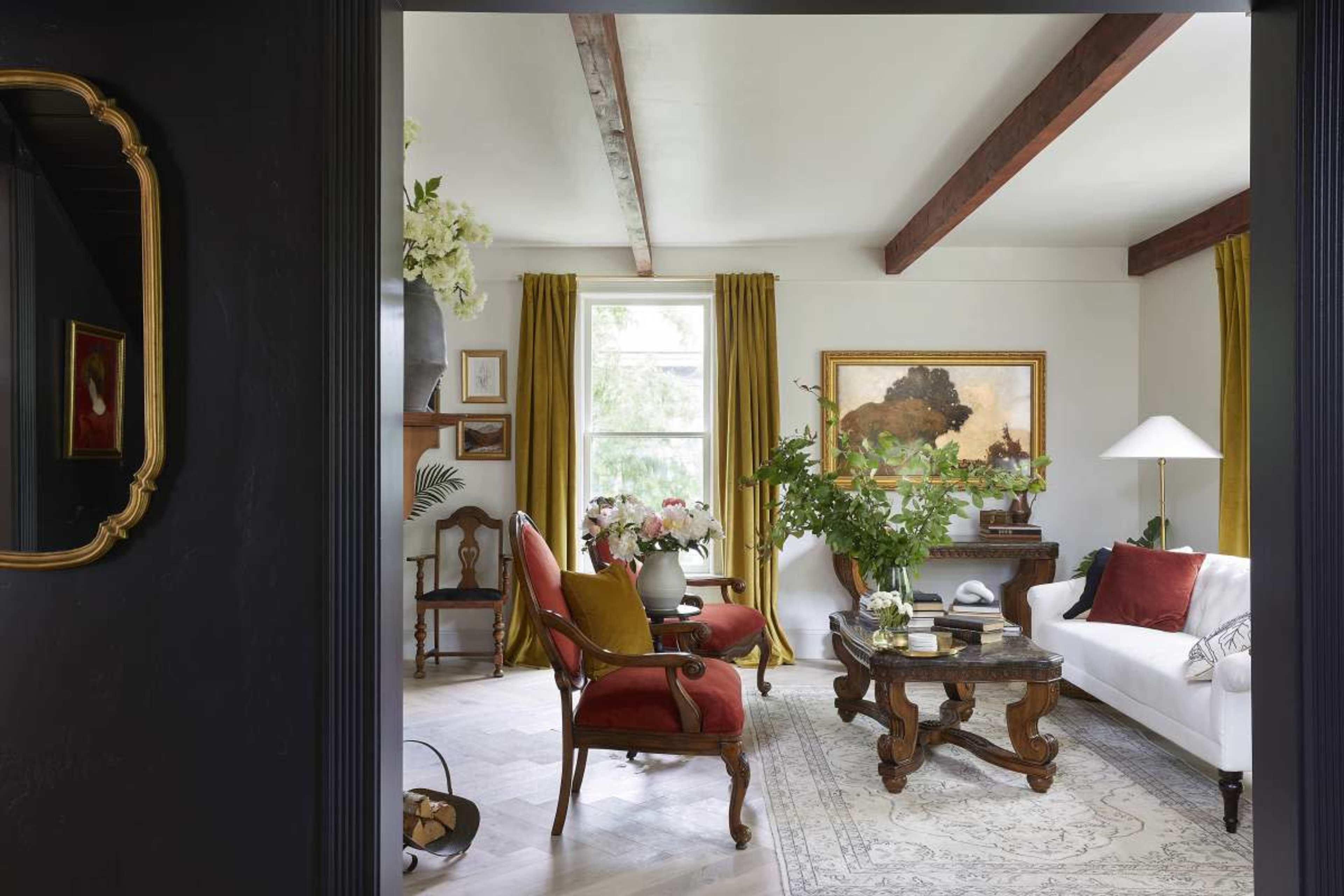 The image shows a living room with a white sofa, a wooden coffee table, and an ornate armchair, featuring large windows dressed with yellow curtains and a decorative painting on the wall.