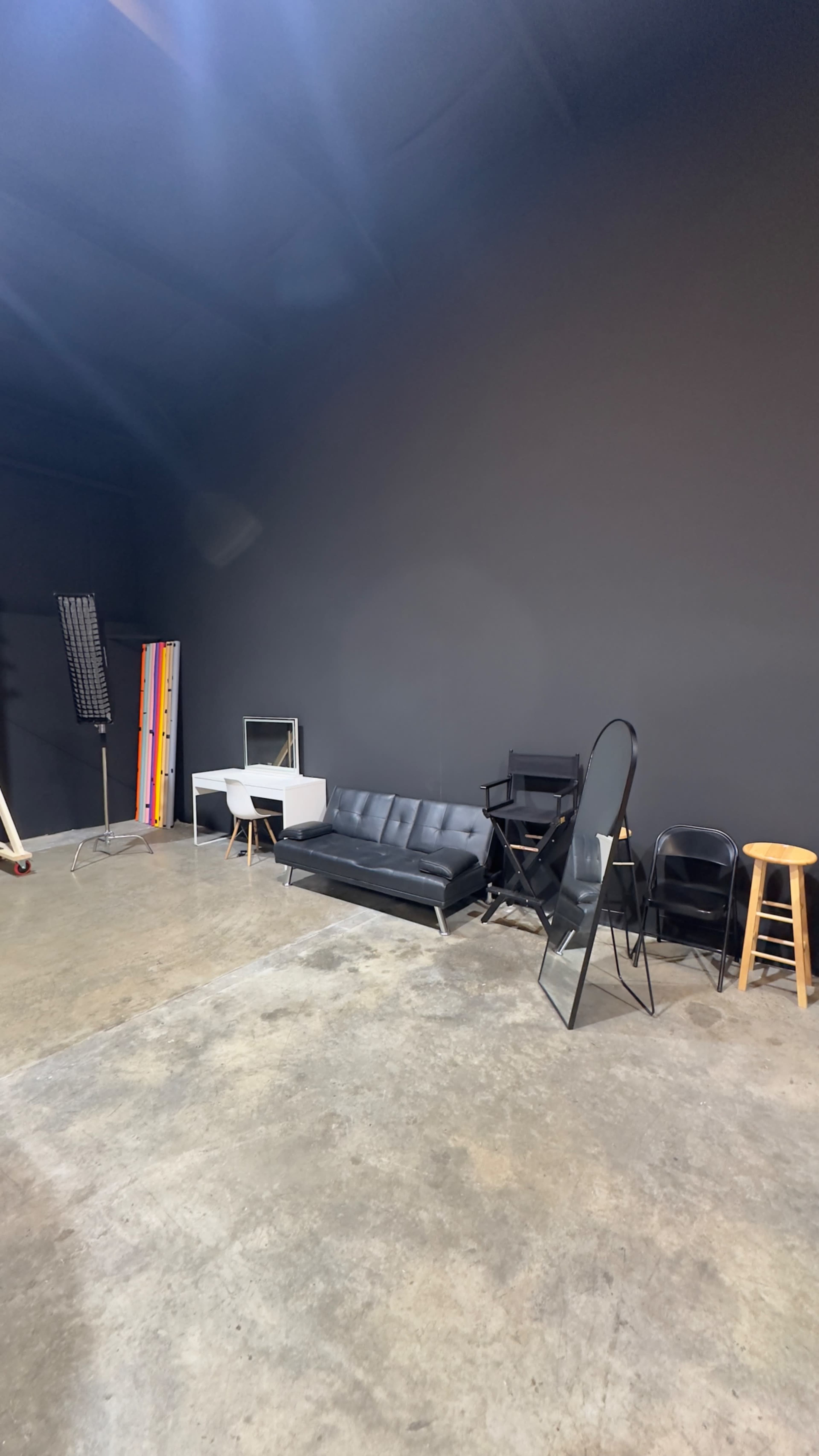 The image shows a spacious studio with a black wall, a black couch, various chairs, a mirror, and colorful backdrop stands.