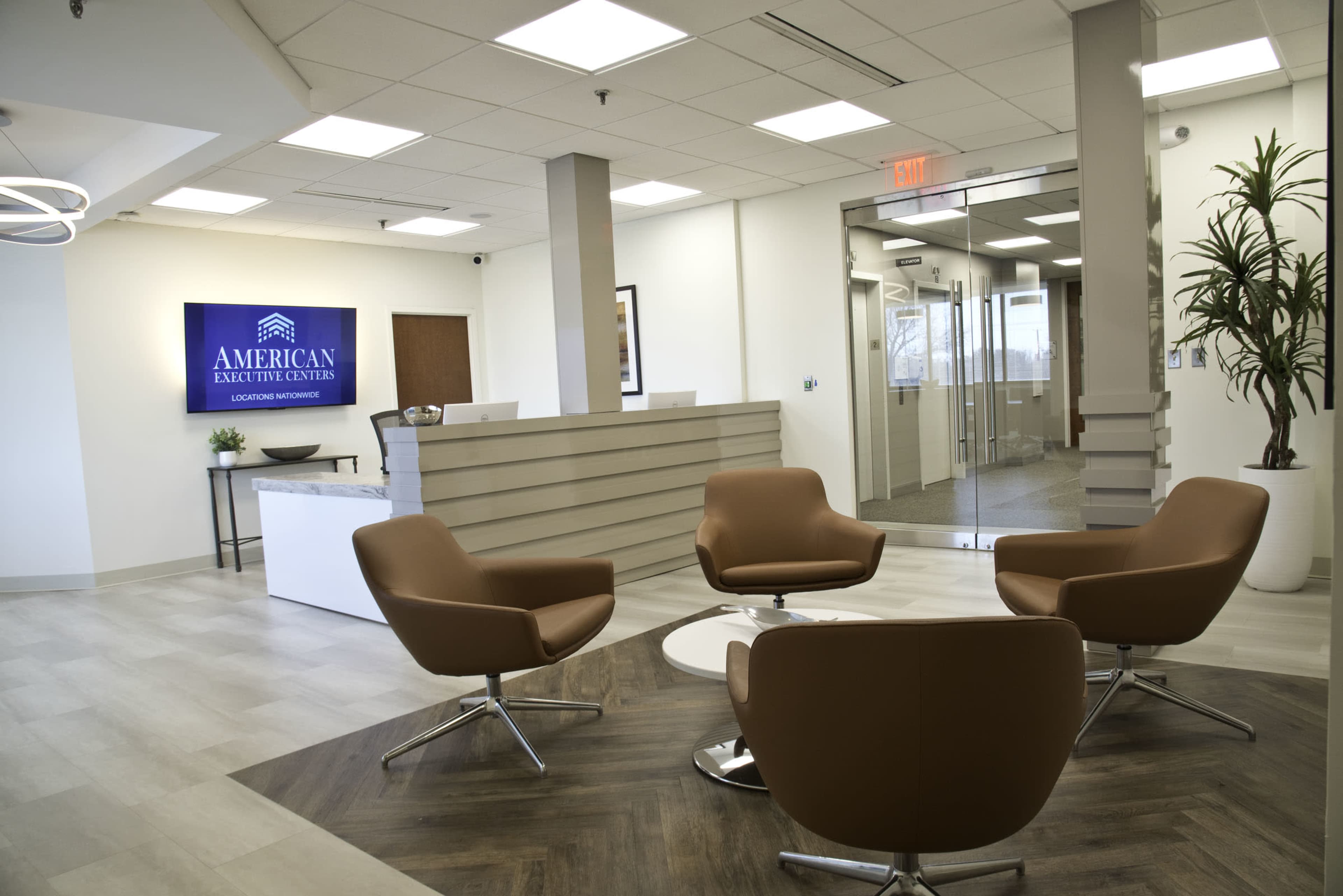 A modern office reception area with a seating arrangement consisting of brown chairs, a circular table, and a large wall-mounted screen displaying the name "American Executive Centers."
