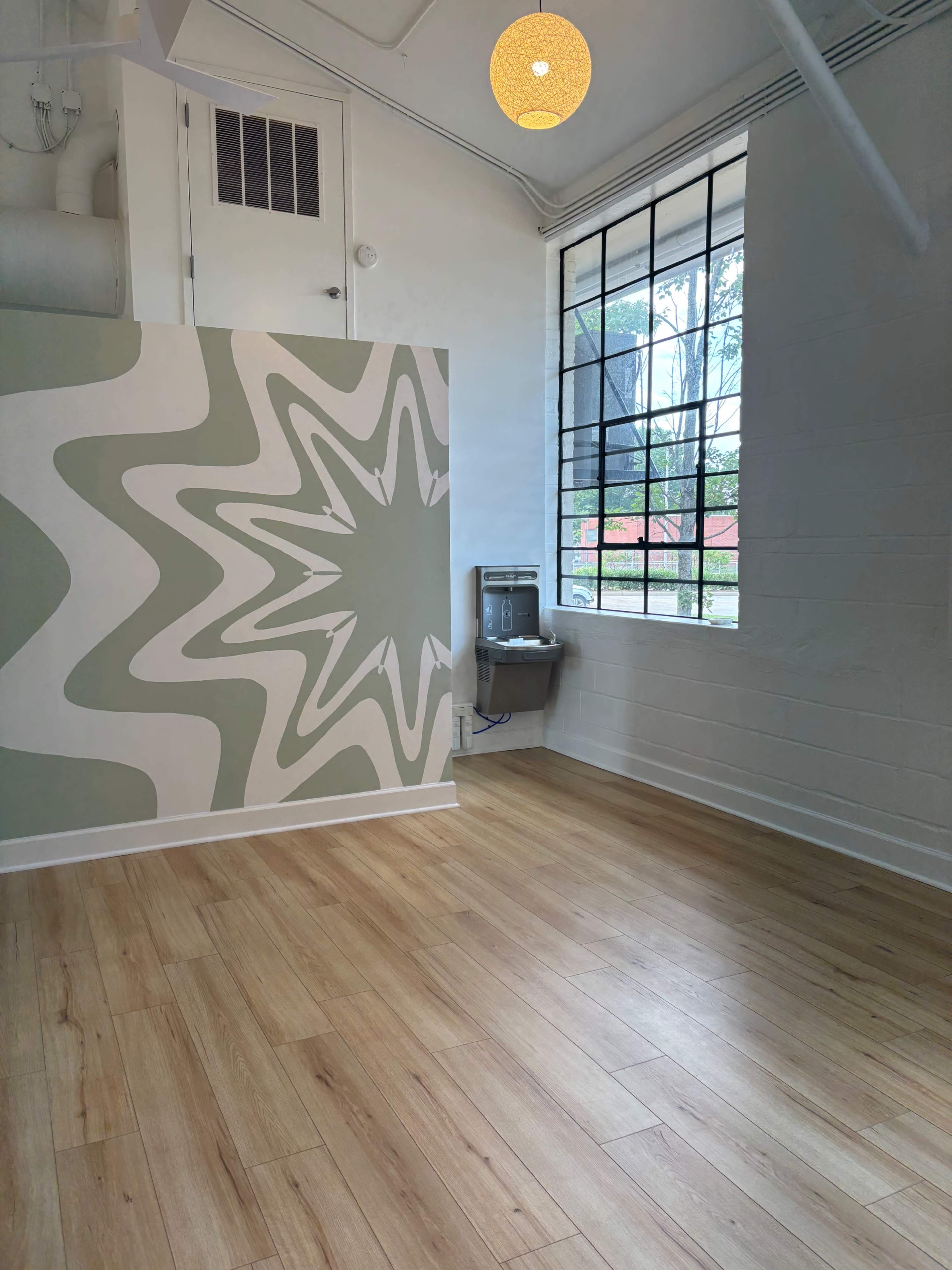 The image shows a spacious room with light wood flooring, a large window, a decorative wall with a starburst pattern, and a drinking water fountain.