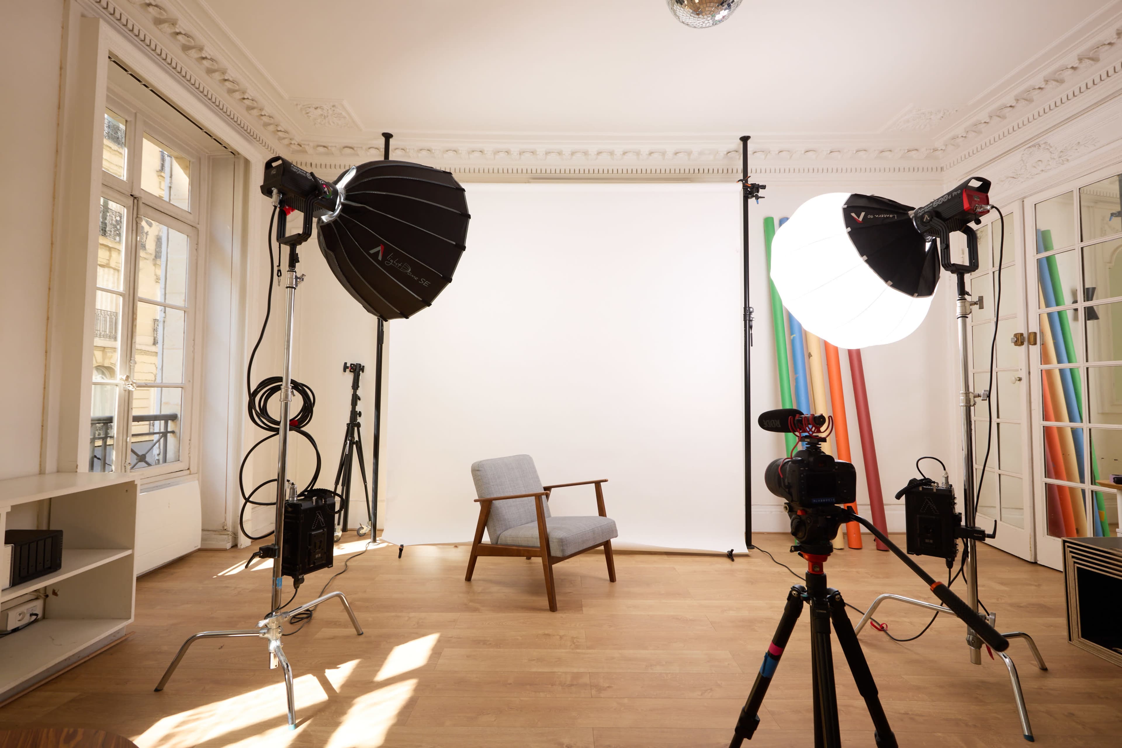 The image shows a photography studio with a gray chair centered in front of a white backdrop, flanked by large softbox lights and camera equipment.