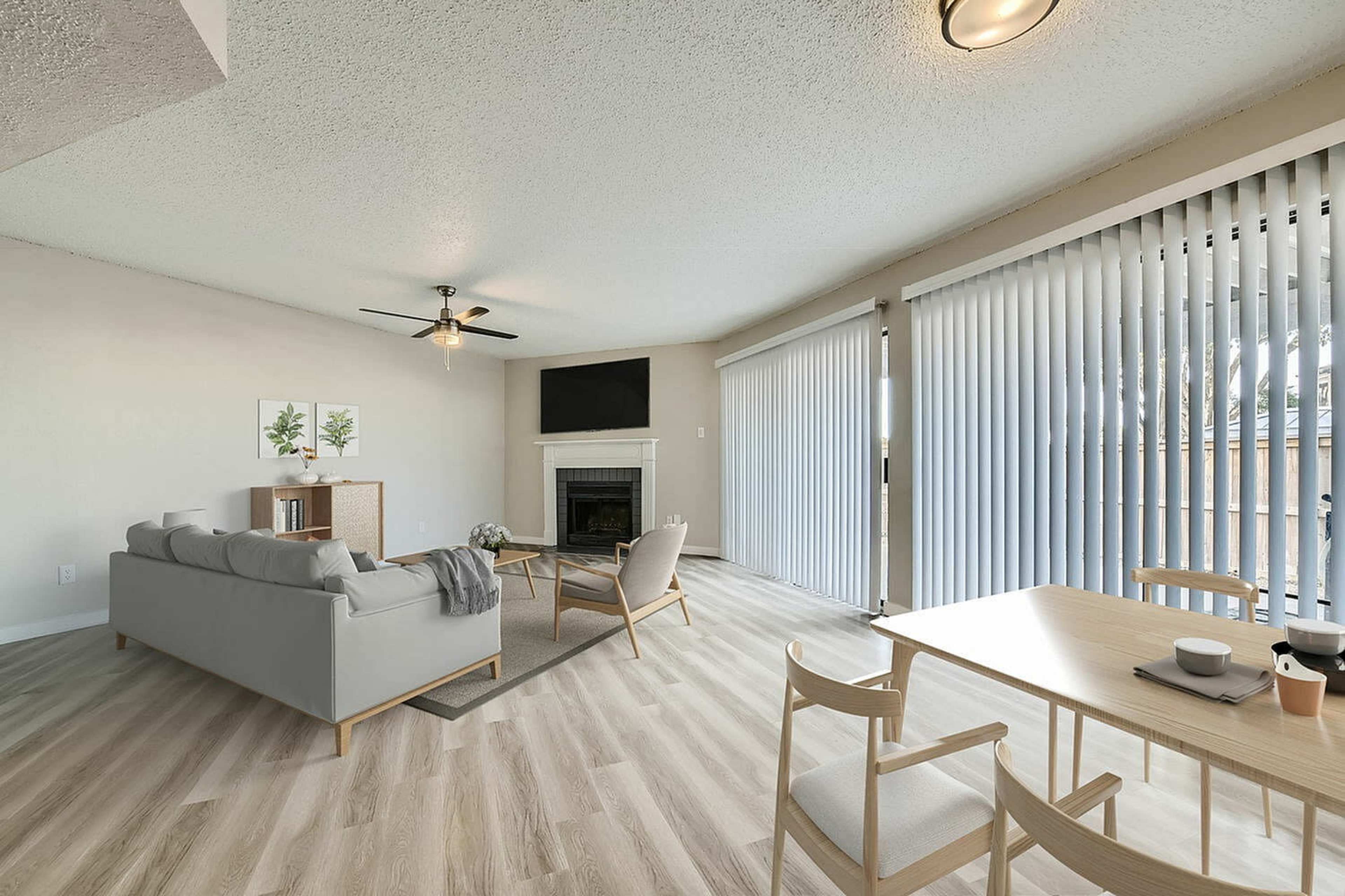 The image shows a modern living room with a gray sofa, wooden dining table, and large windows covered by vertical blinds.