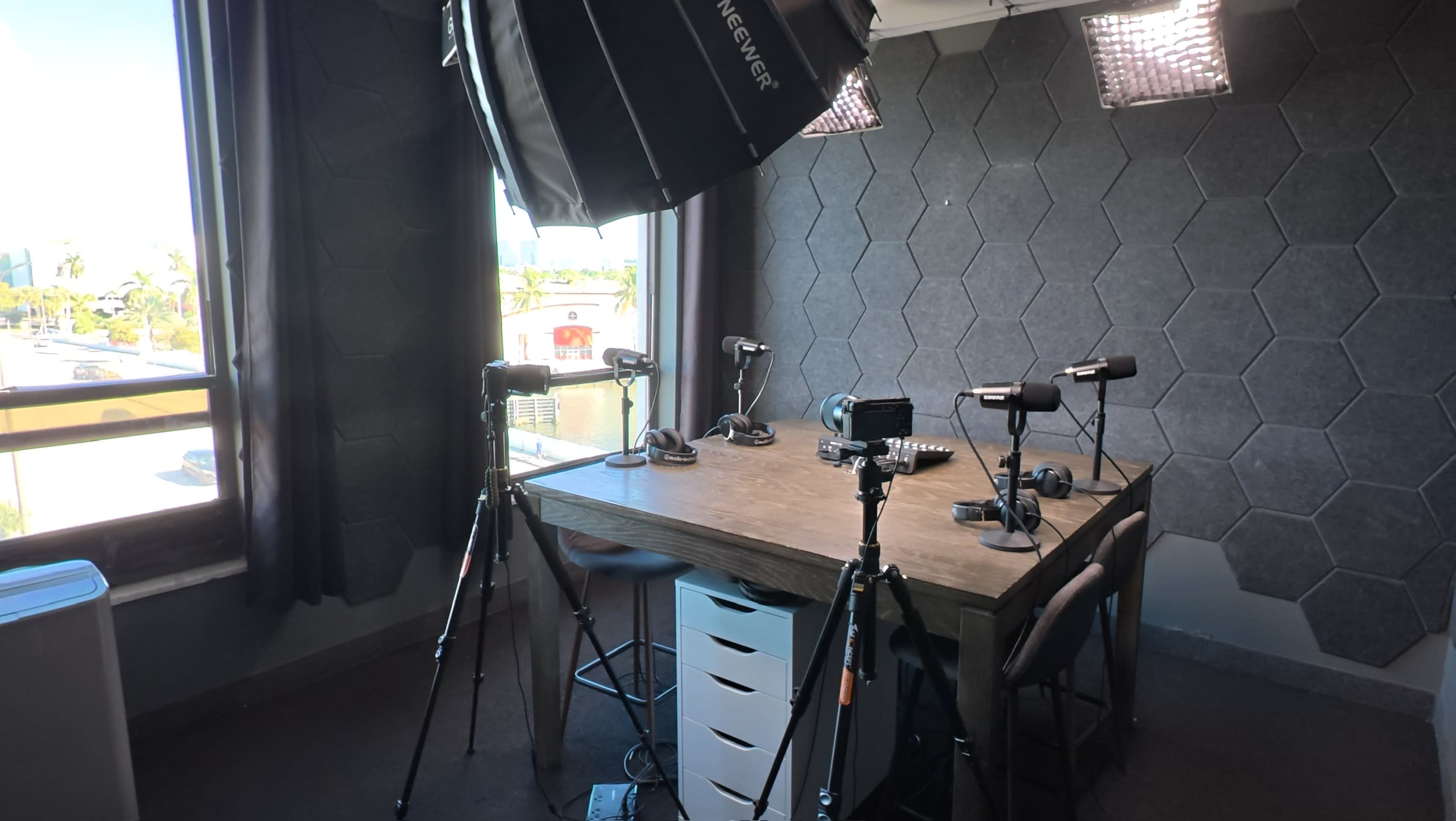 A well-lit recording studio features a hexagon-patterned wall, a wooden table, and multiple cameras and microphones set up for a video production.