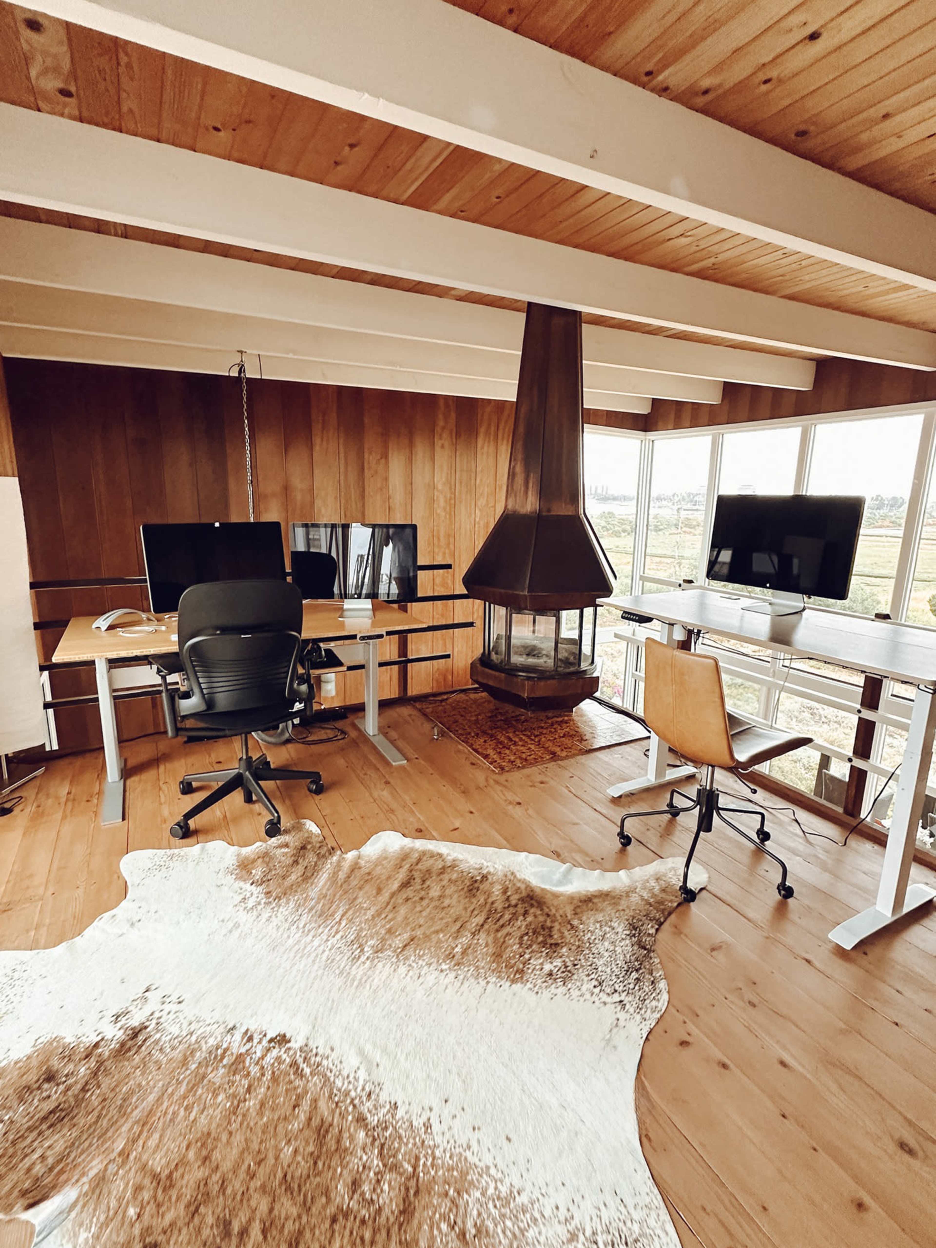 A modern workspace featuring two computer desks, a swivel chair, a fireplace, and a cowhide rug on a wooden floor.