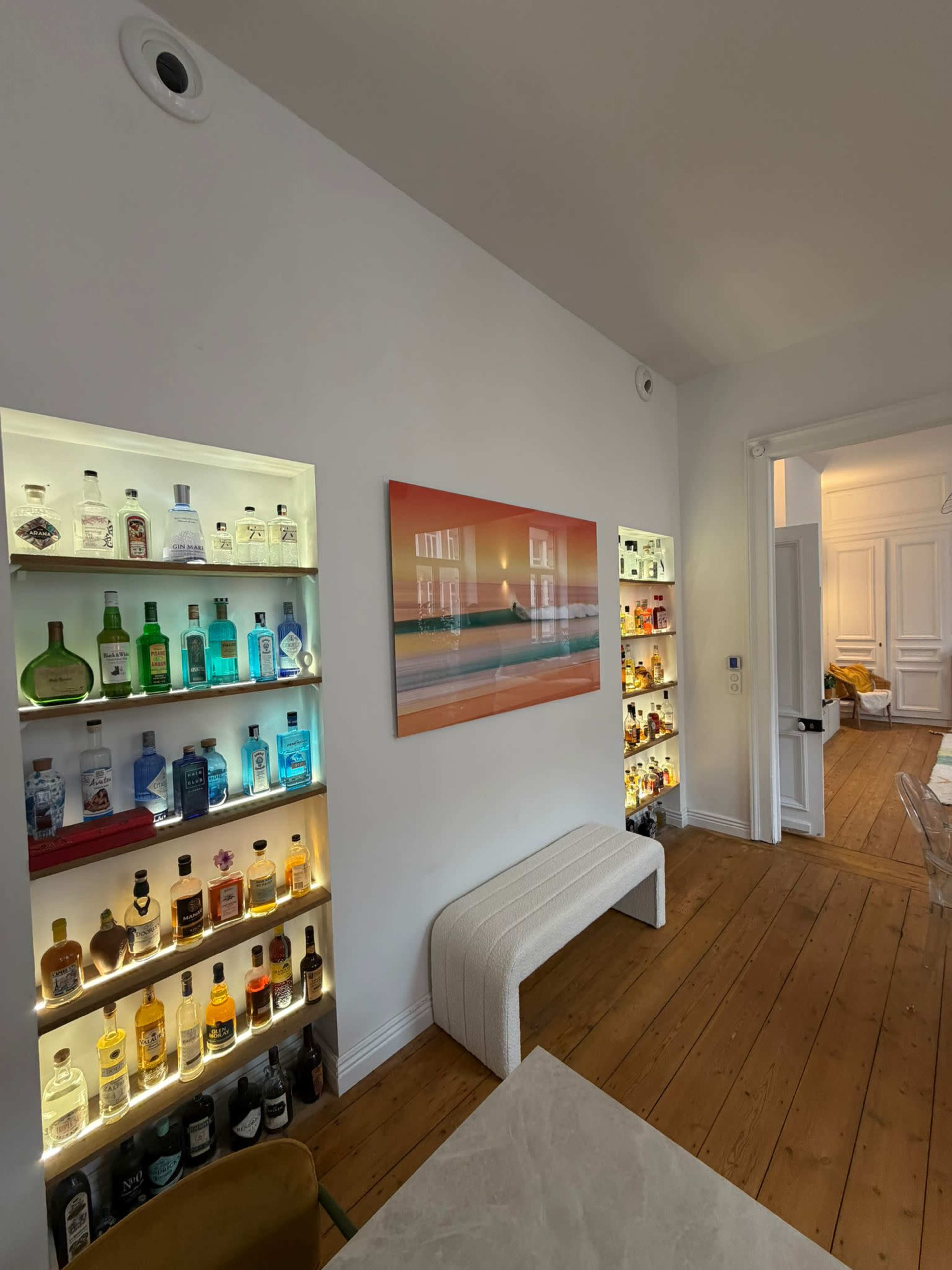 The image shows a well-lit room with two illuminated shelving units displaying various bottles of alcohol, a minimalist bench, and a colorful painting on the wall opposite a doorway.