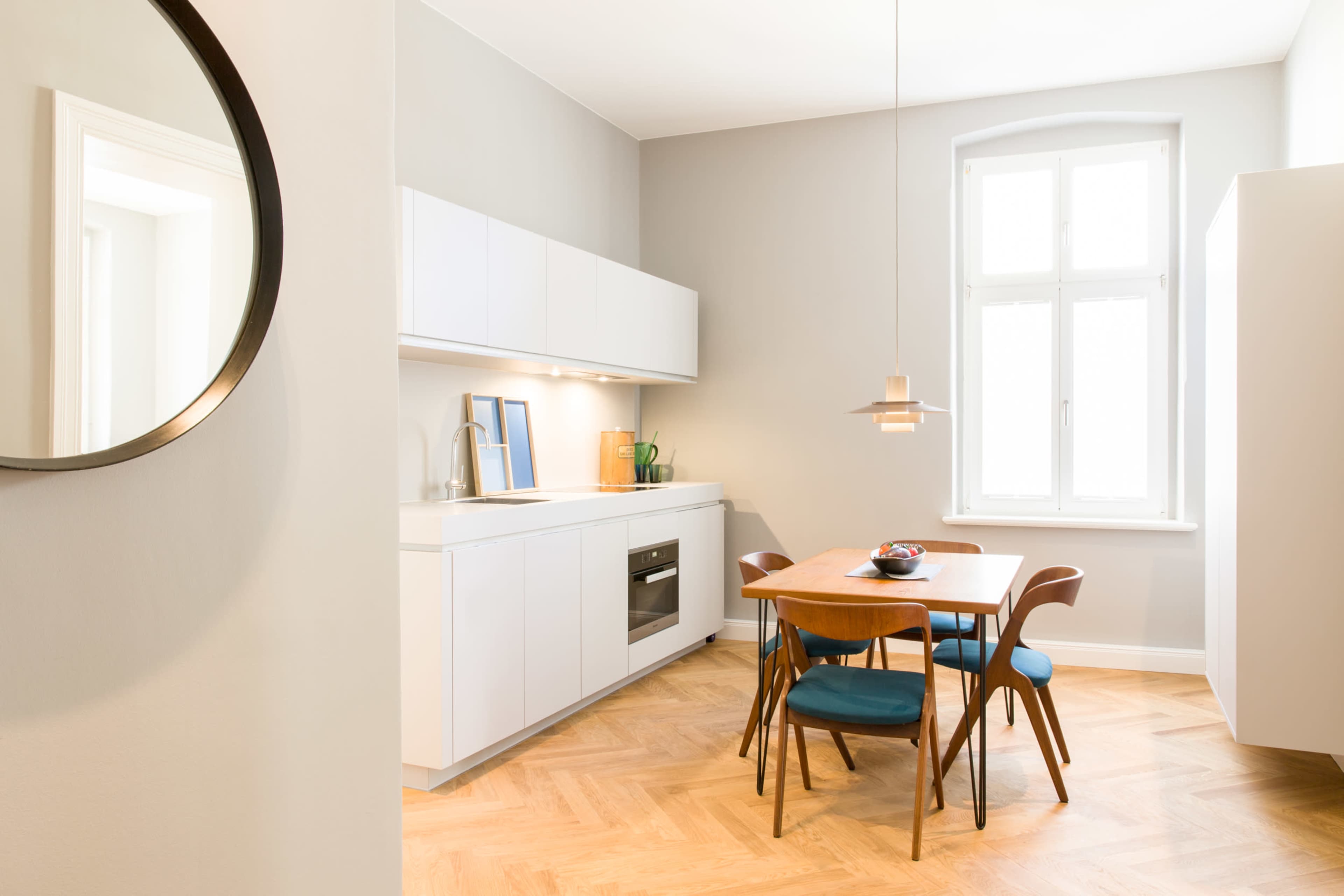 A modern kitchen with a wooden dining table, four chairs, sleek white cabinetry, and large windows allowing natural light.