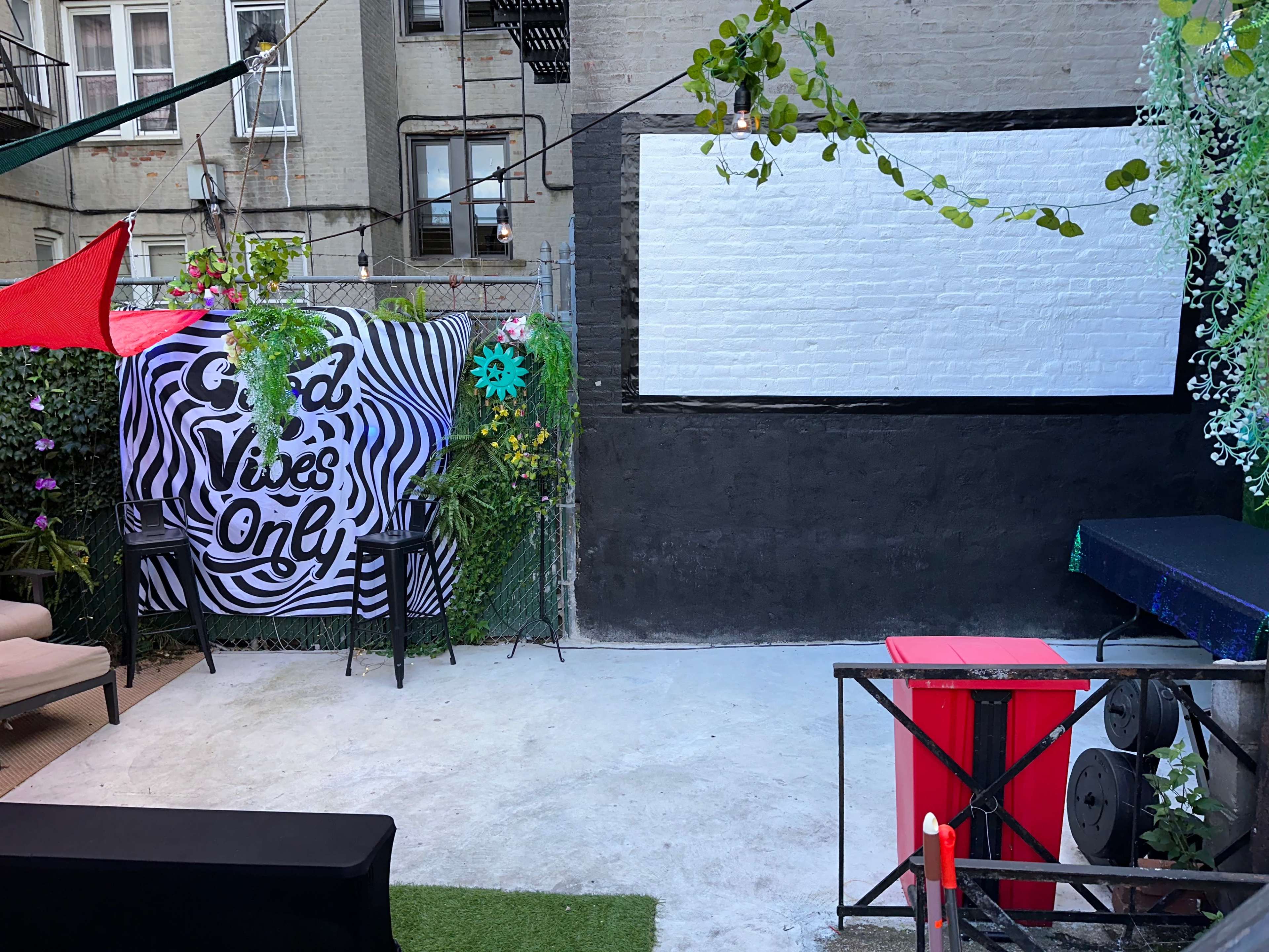 An outdoor space with a large white screen mounted on a wall, surrounded by greenery and a decorative backdrop that reads "Good Vibes Only."