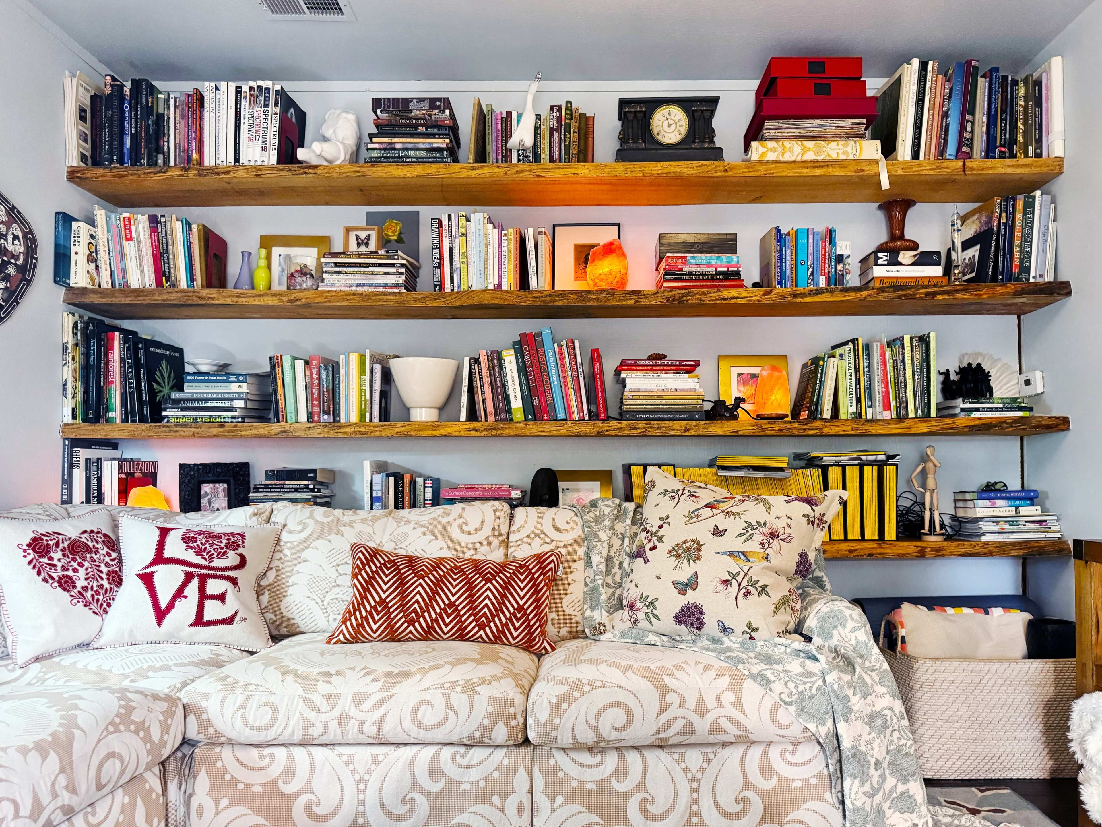 A cozy living room features a light-colored sofa adorned with decorative pillows, in front of a wall-mounted bookshelf filled with books and various decor items.