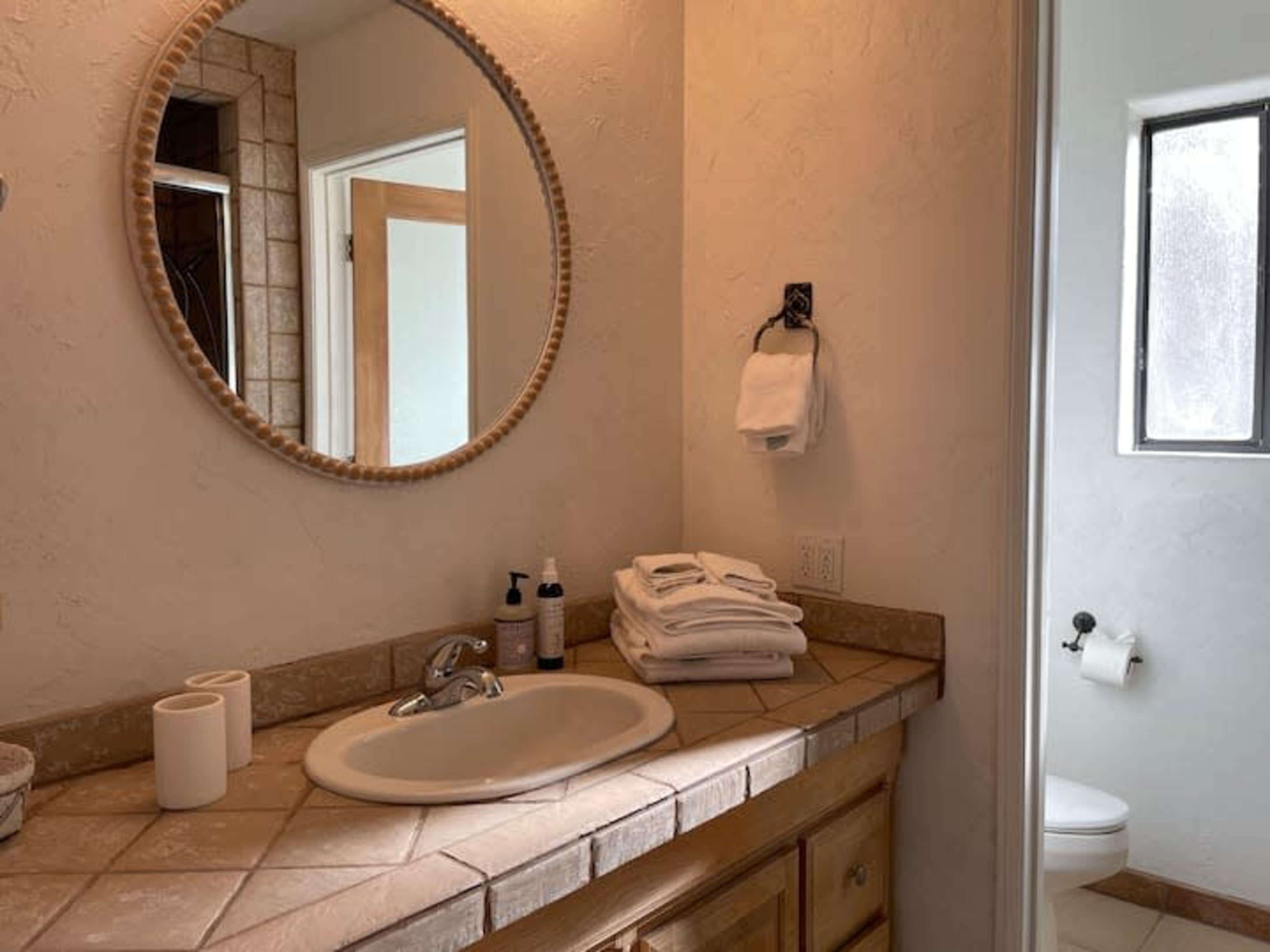 A bathroom with a round mirror above a sink, a neatly arranged stack of white towels, and a door leading to a room with a toilet.