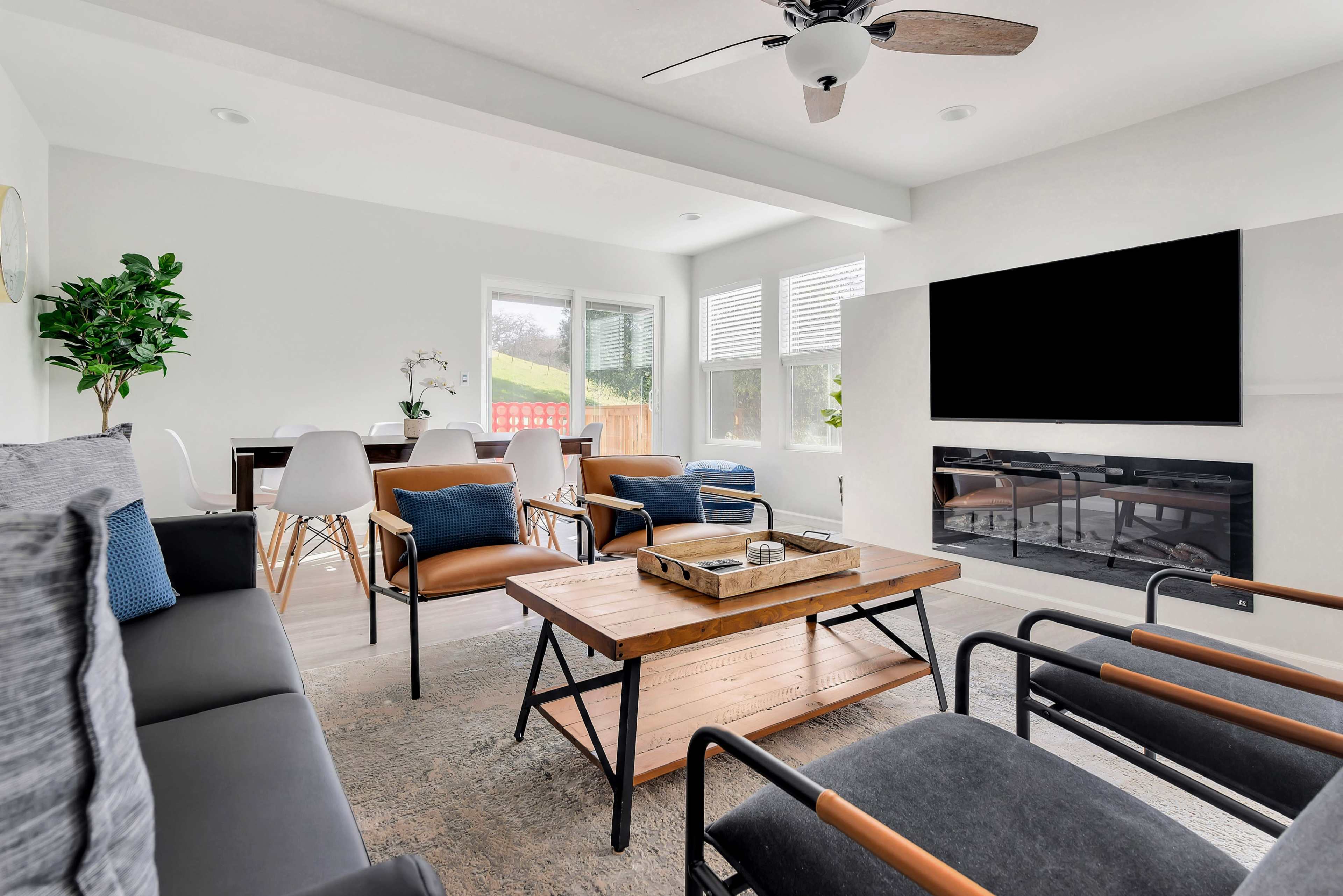 A modern living room features a central wooden coffee table surrounded by various seating options and a wall-mounted TV.