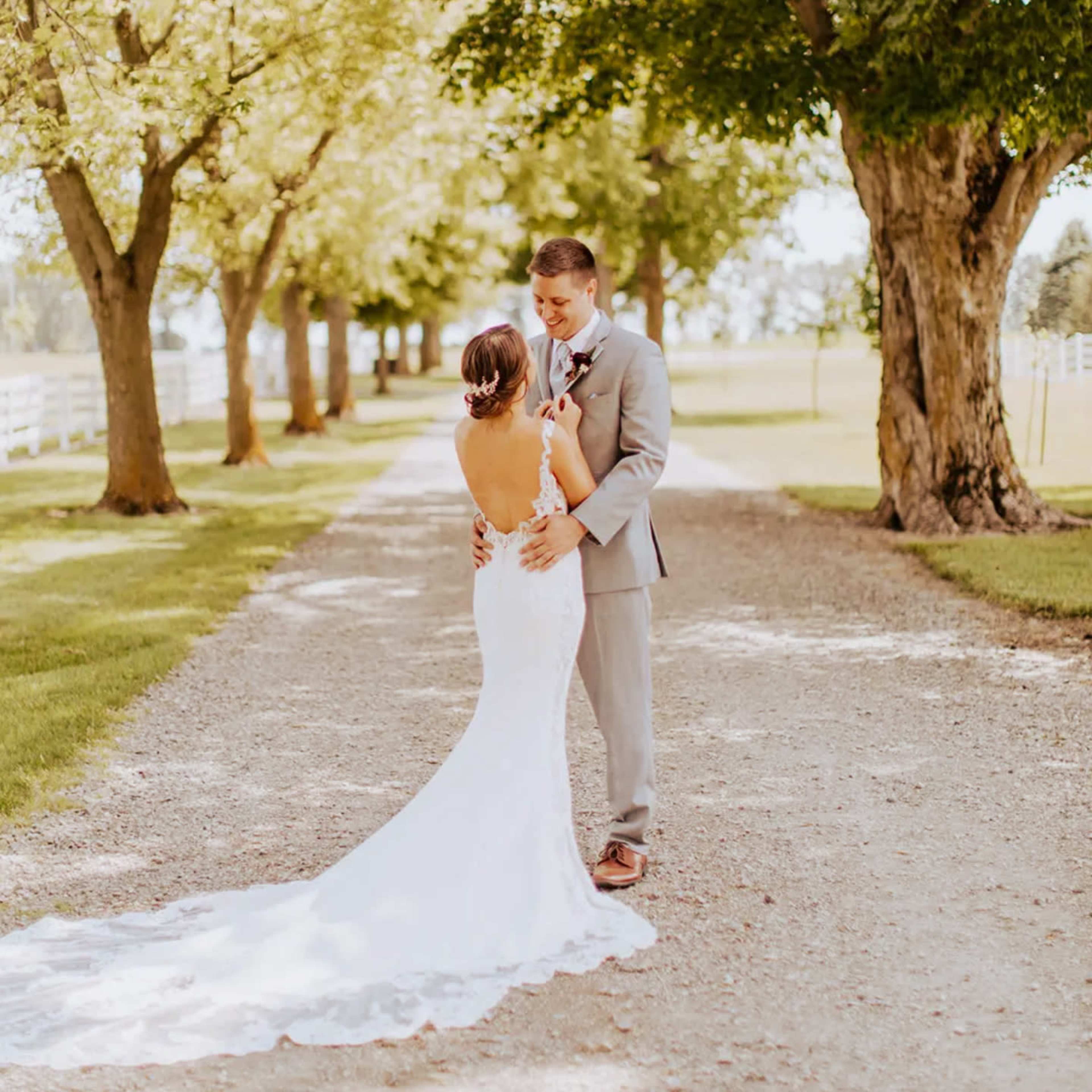 A couple stands together on a gravel path lined with trees, with the woman in a white wedding dress and the man in a gray suit.