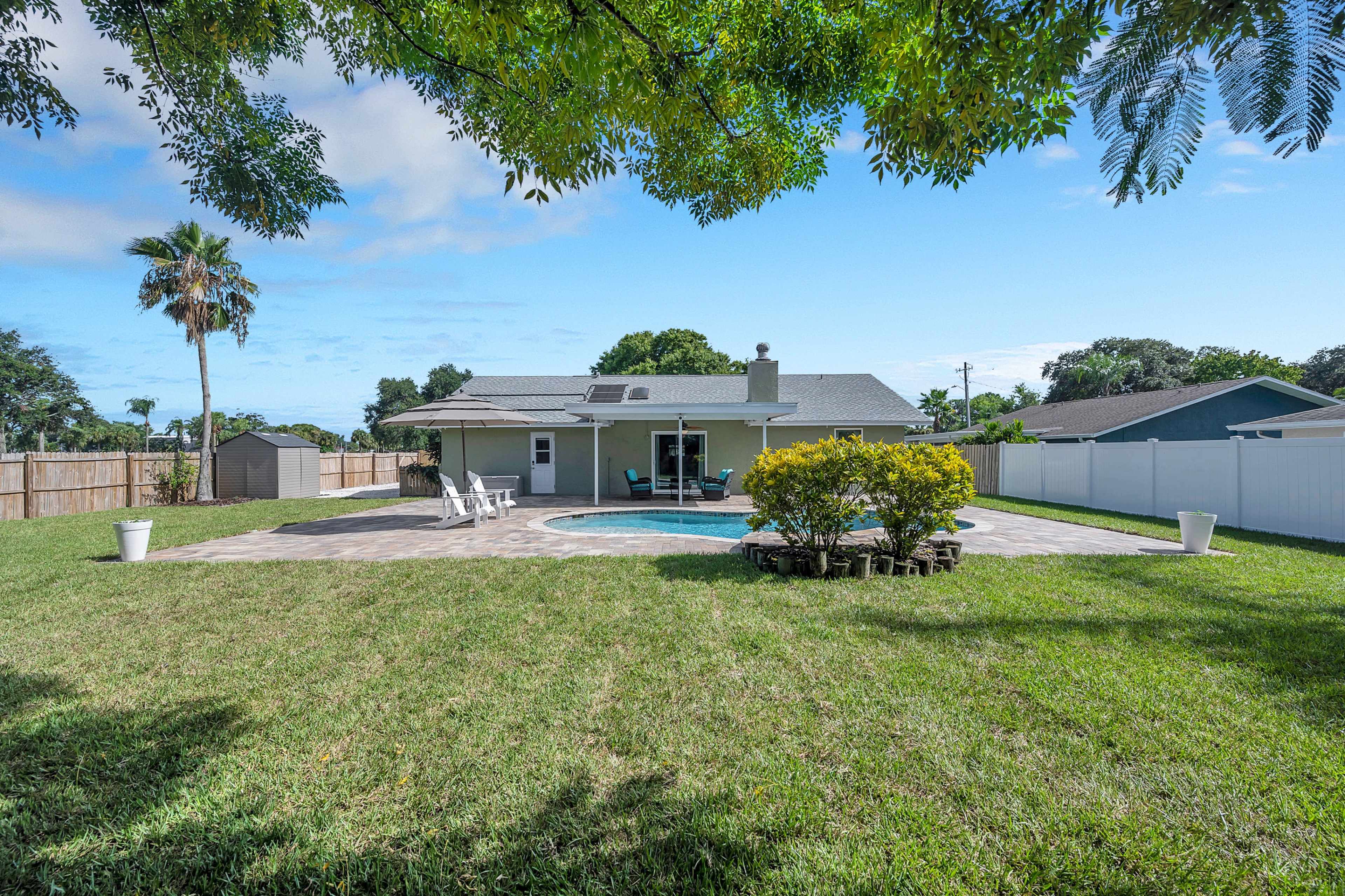The image shows a backyard with a swimming pool, patio furniture under an umbrella, and a well-maintained lawn surrounded by a white fence.