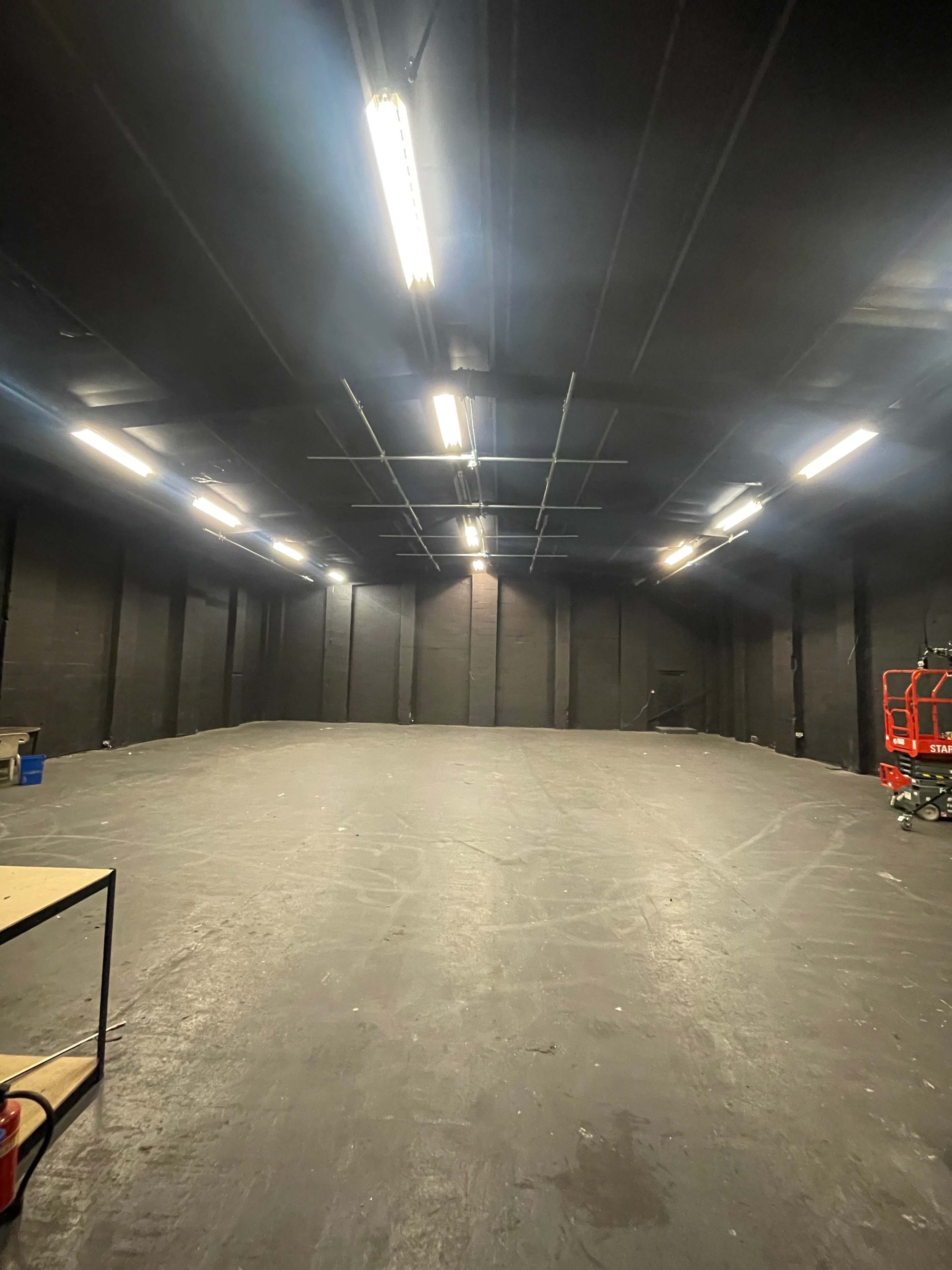 An empty, dimly lit industrial space with black walls and a smooth floor, featuring overhead lights and a small table on the left.