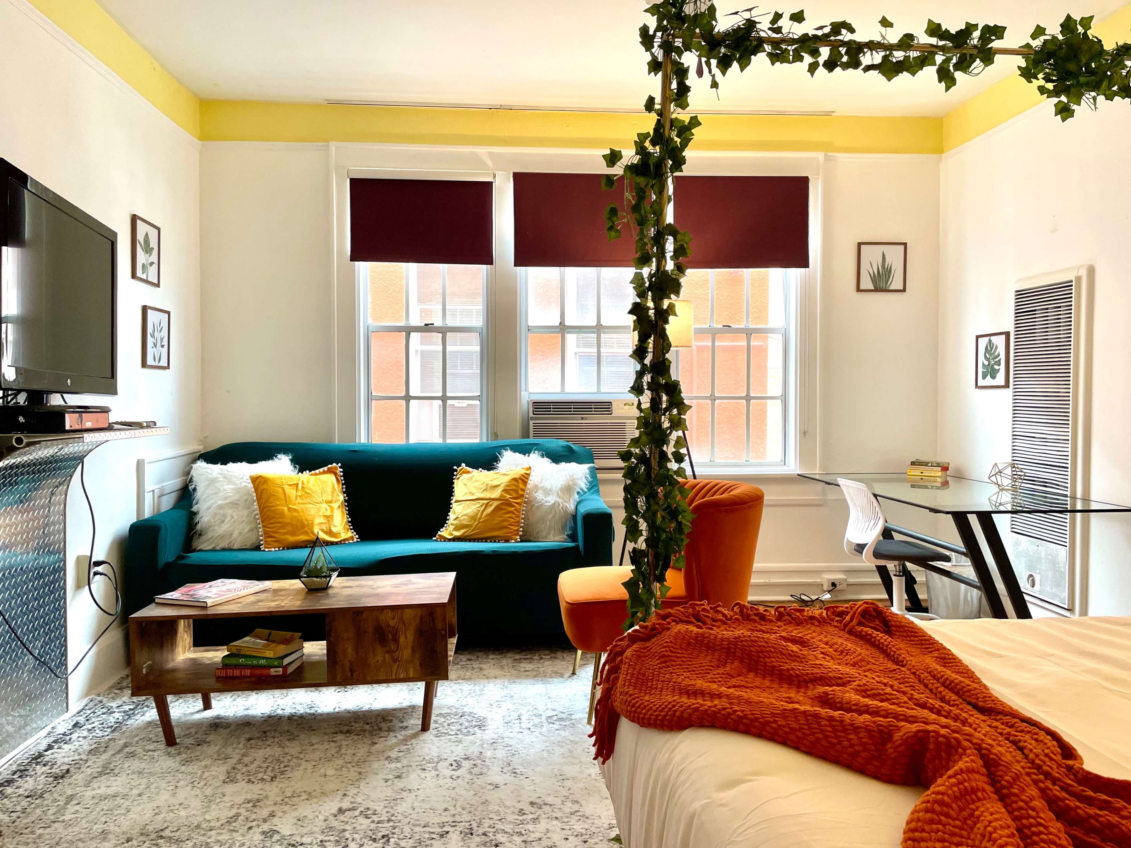 A cozy living space featuring a green couch with yellow pillows, a wooden coffee table, a work desk, and a bed with an orange throw blanket.