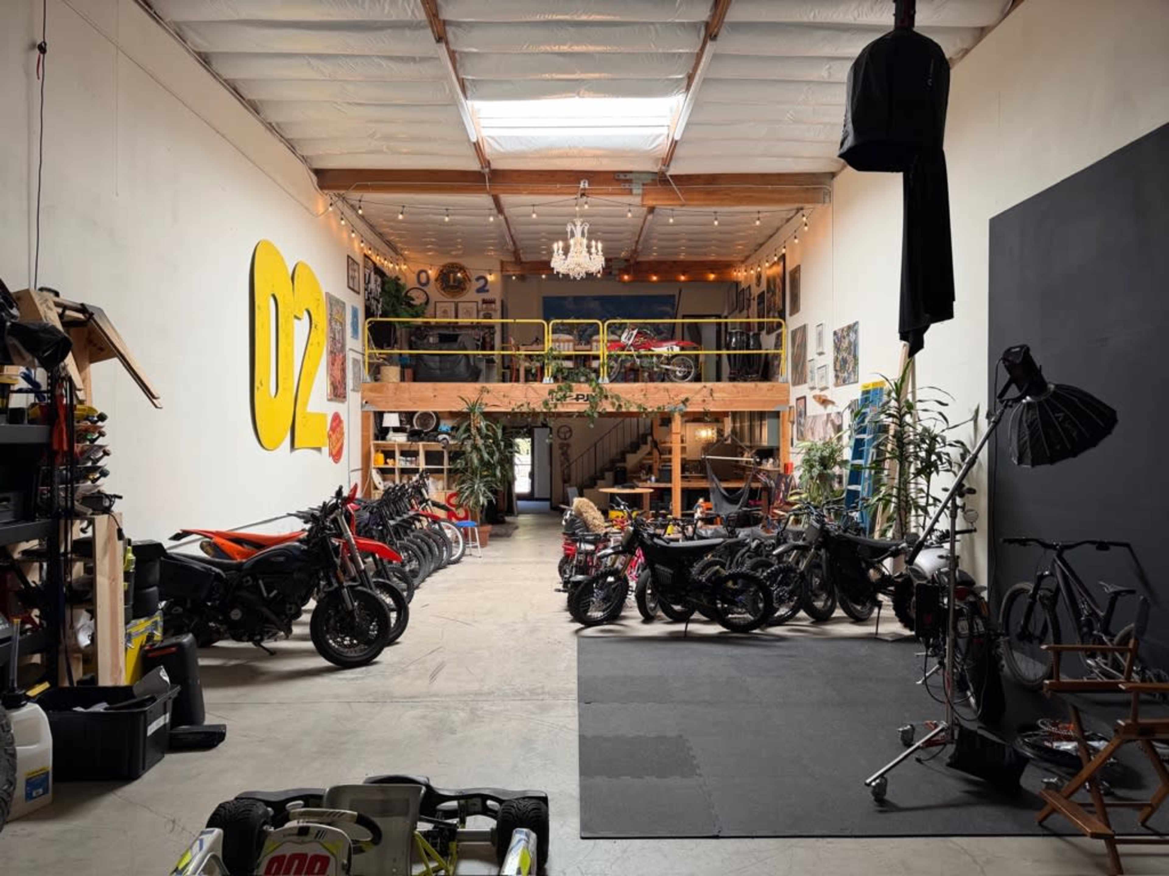 The image shows a spacious garage with various motorcycles lined up, a mezzanine level adorned with plants and artwork, and lighting that creates an inviting atmosphere.