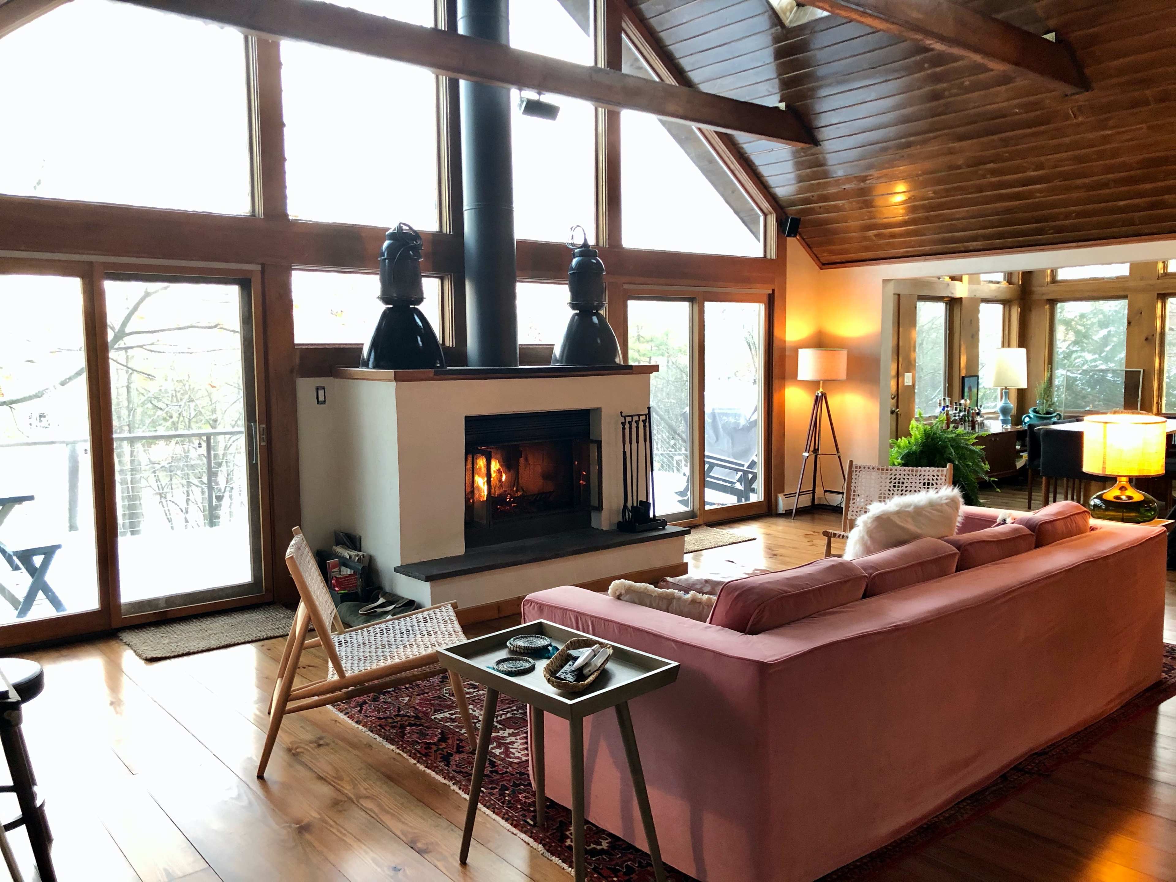 A cozy living room with a pink sofa facing a fireplace, large windows letting in natural light, and wooden beams on the ceiling.