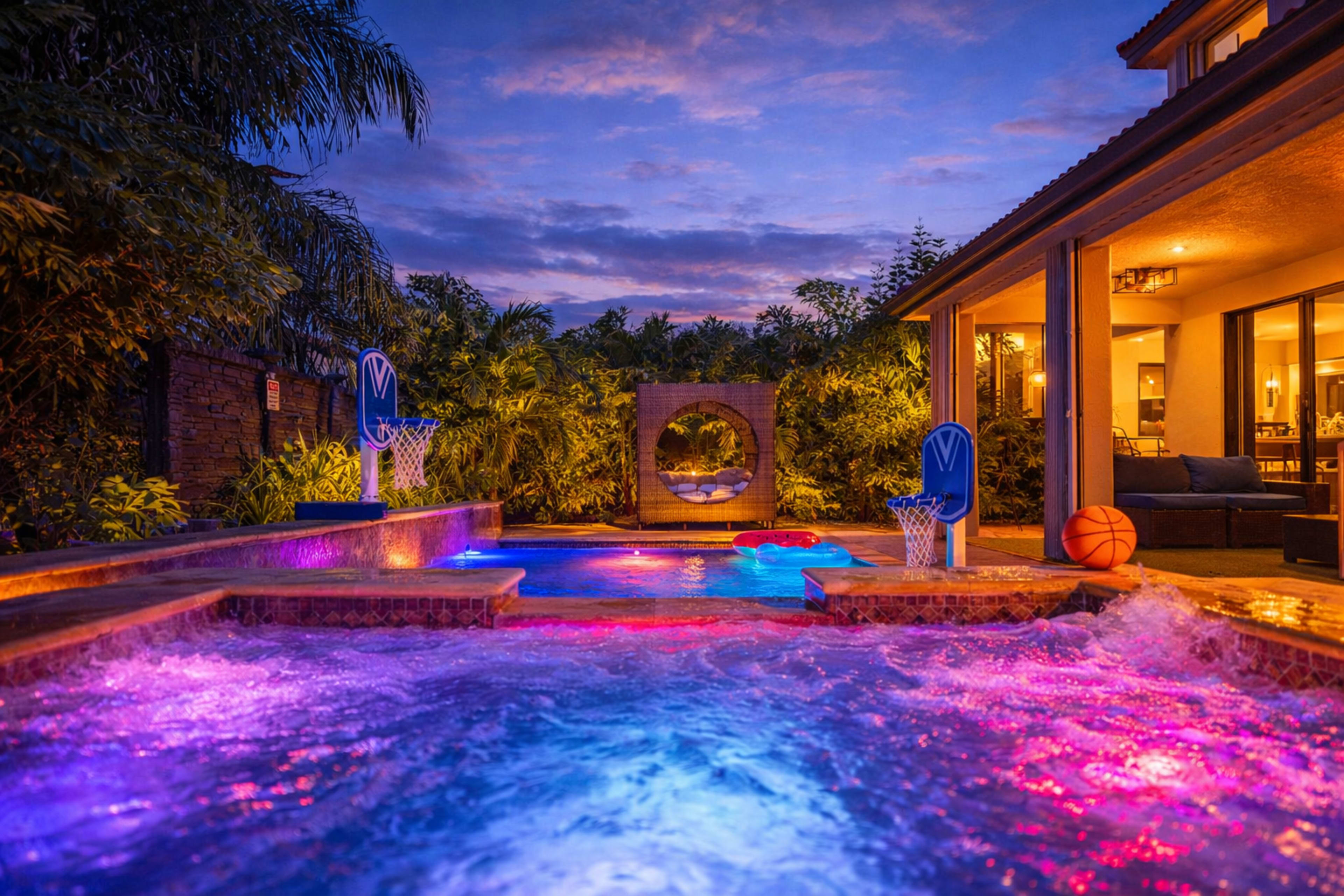A backyard pool area features colorful lights, basketball hoops, and a view of a cozy seating space in the background, all set against a twilight sky.