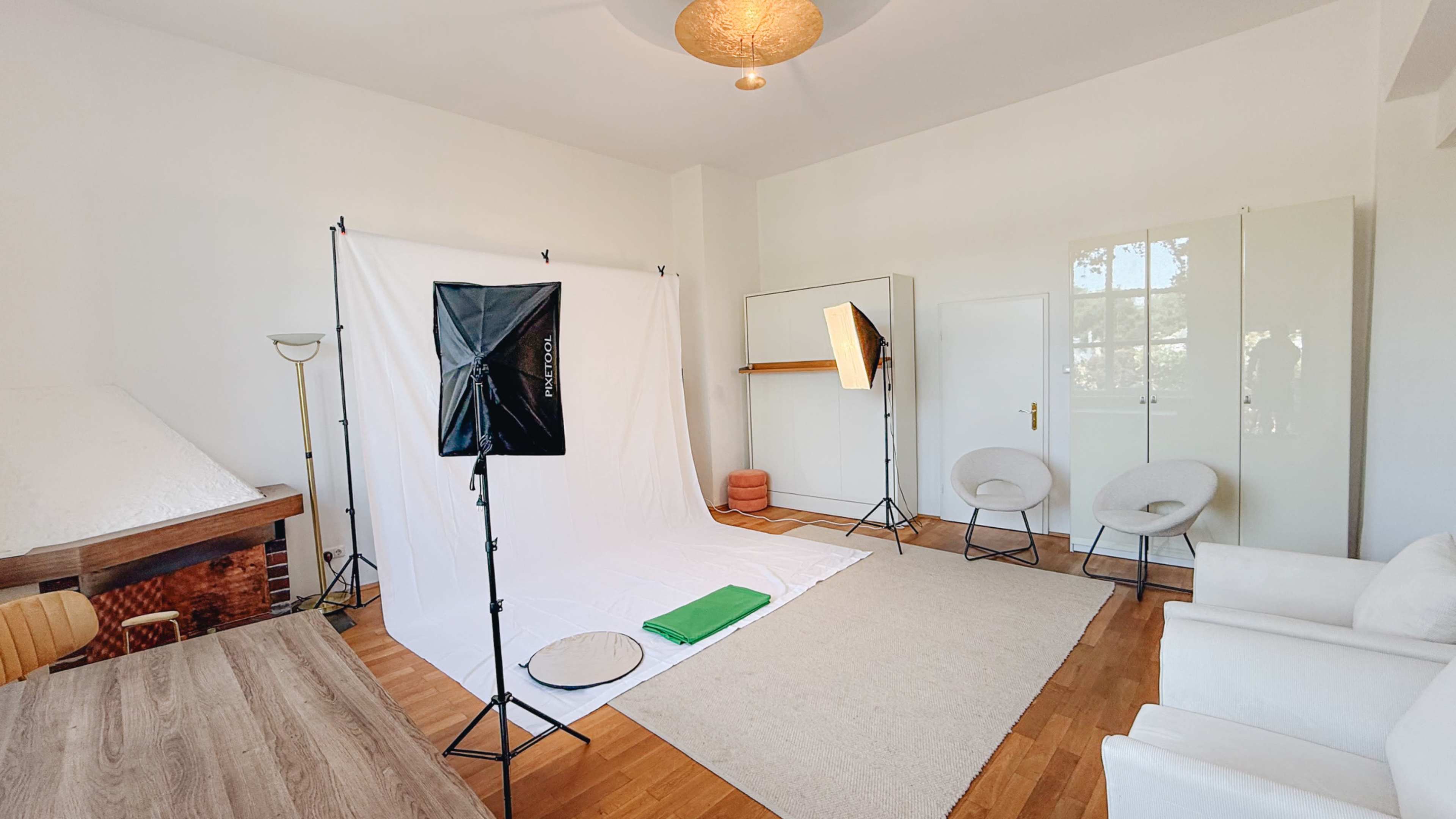 The image shows a well-lit photography studio featuring a white backdrop, two light stands, a rug, and several pieces of furniture, including chairs and a table.