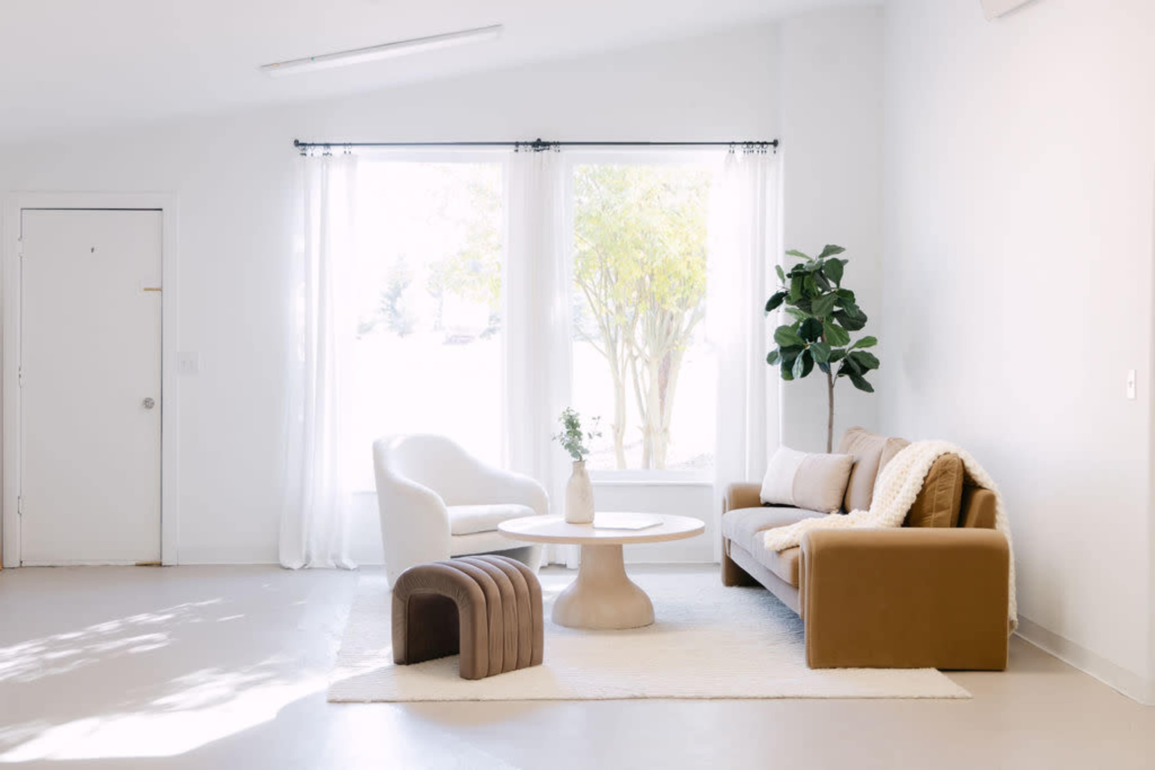 The image shows a modern living room with a white chair, a brown sofa, a round coffee table, and a large window with sheer curtains, allowing natural light to enter.