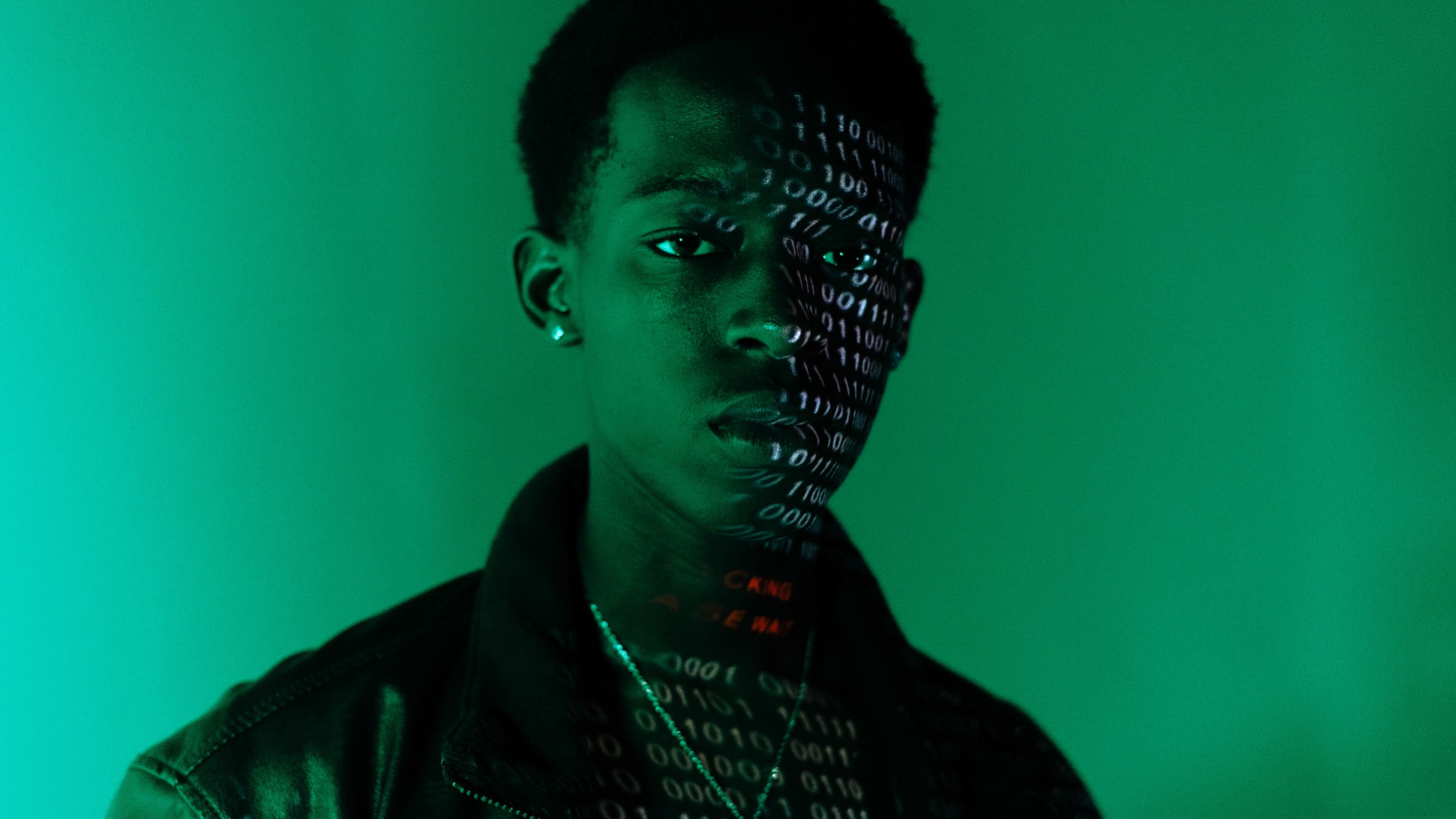 A young person stands against a green backdrop with binary code projected onto half of their face.