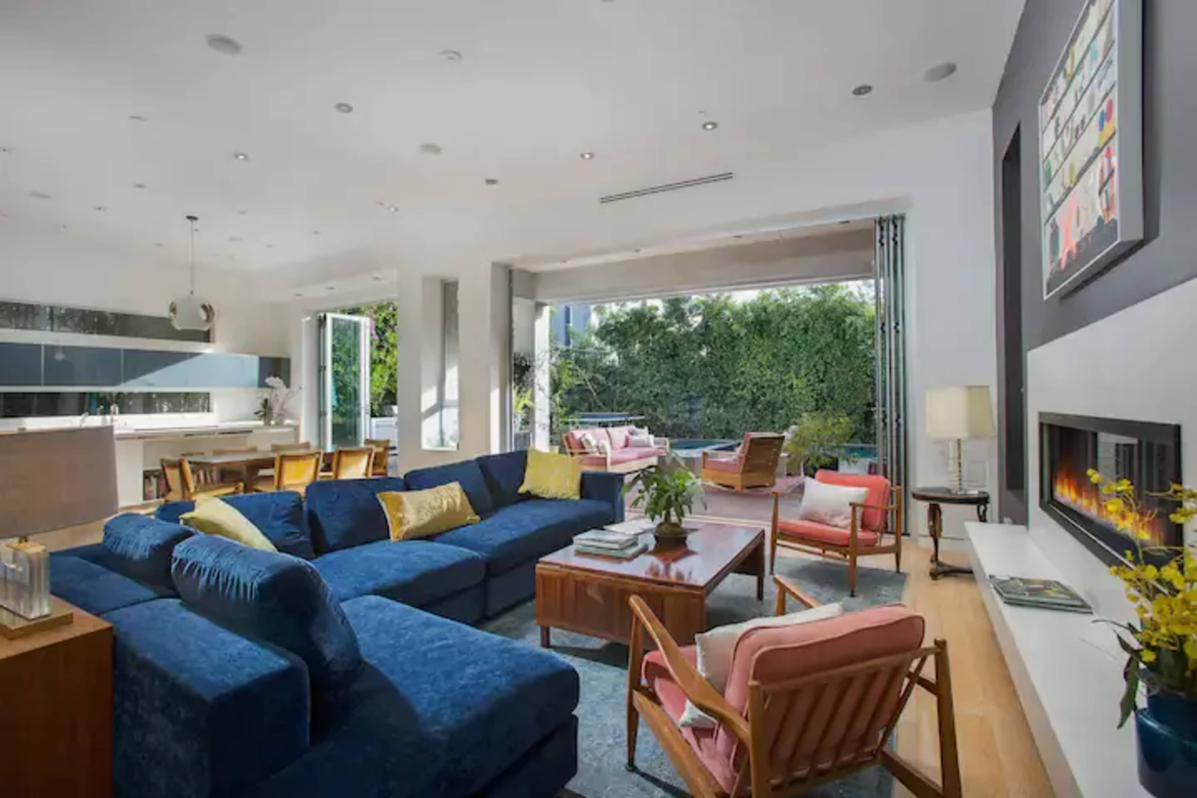 A modern living room features a large blue sectional sofa, wooden furniture, and an open layout leading to an outdoor space.