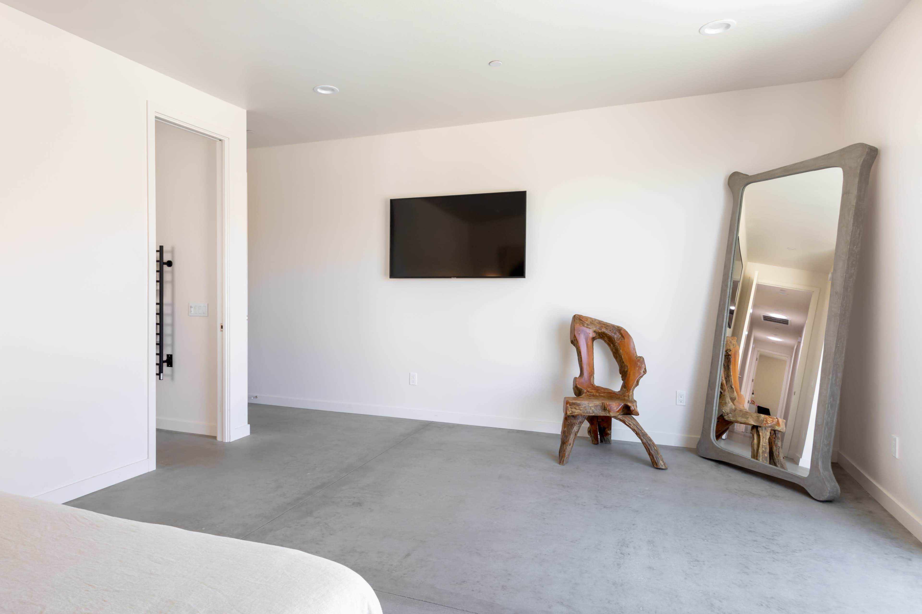 A minimalist bedroom features a wall-mounted flat-screen TV, a large mirror, and a unique wooden chair on a concrete floor.