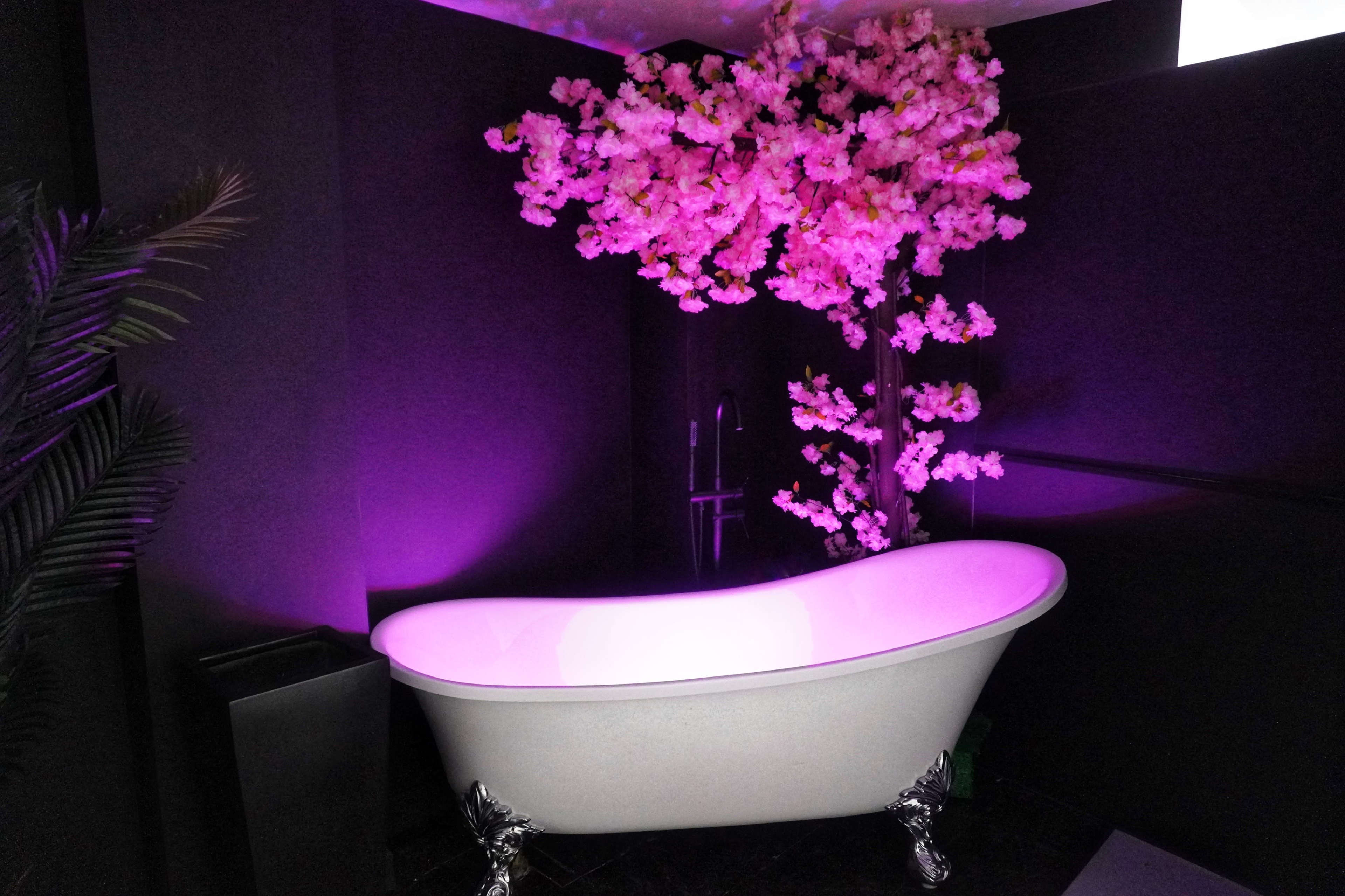 A freestanding bathtub is positioned in a dimly lit room adorned with pink flowers and purple lighting.