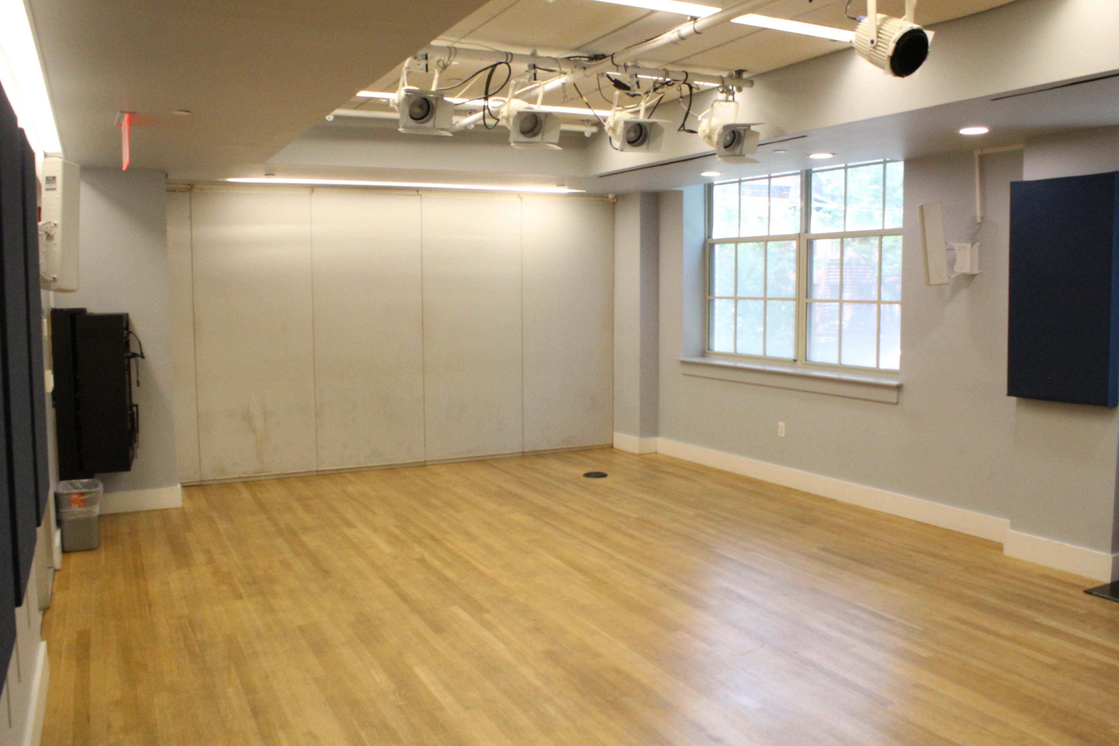 The image shows an empty room with wooden flooring, gray walls, a large window on one side, and ceiling-mounted lights.
