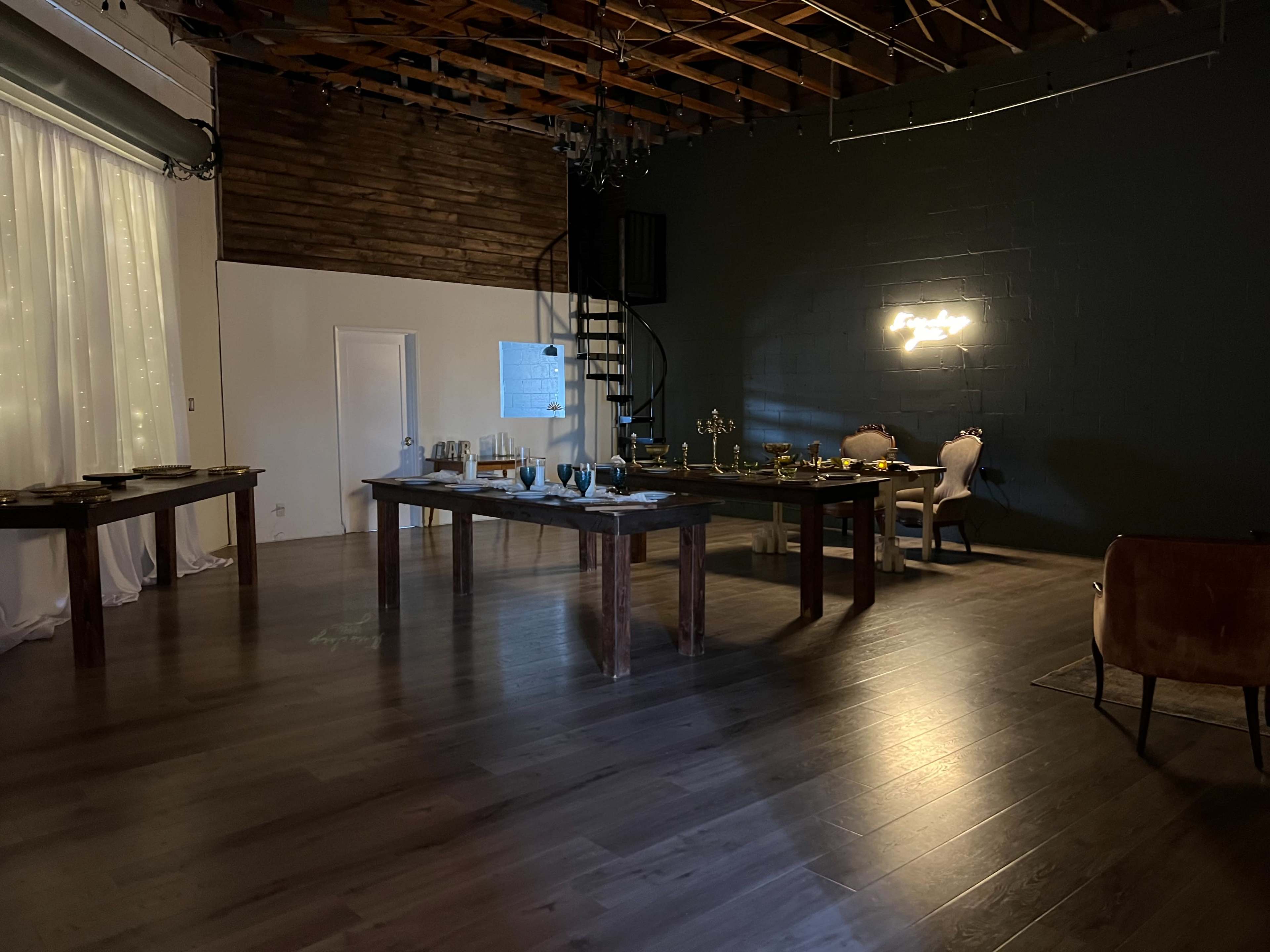 A spacious, dimly lit room with wooden tables arranged for an event, a staircase in the background, and decorative lighting on the walls.