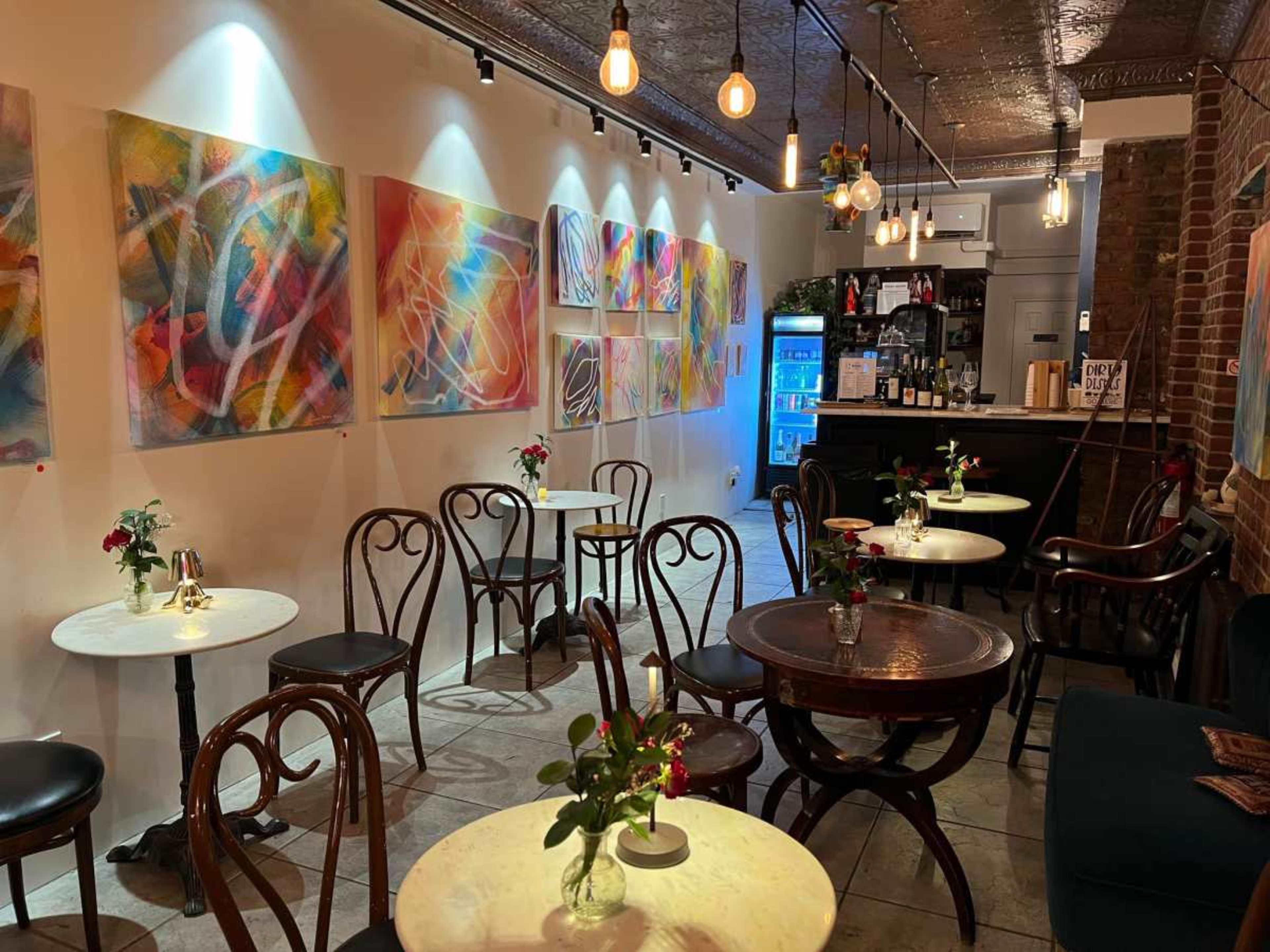 A cozy cafe features several small round tables with flowers, surrounded by colorful abstract paintings on the walls and a bar in the background.