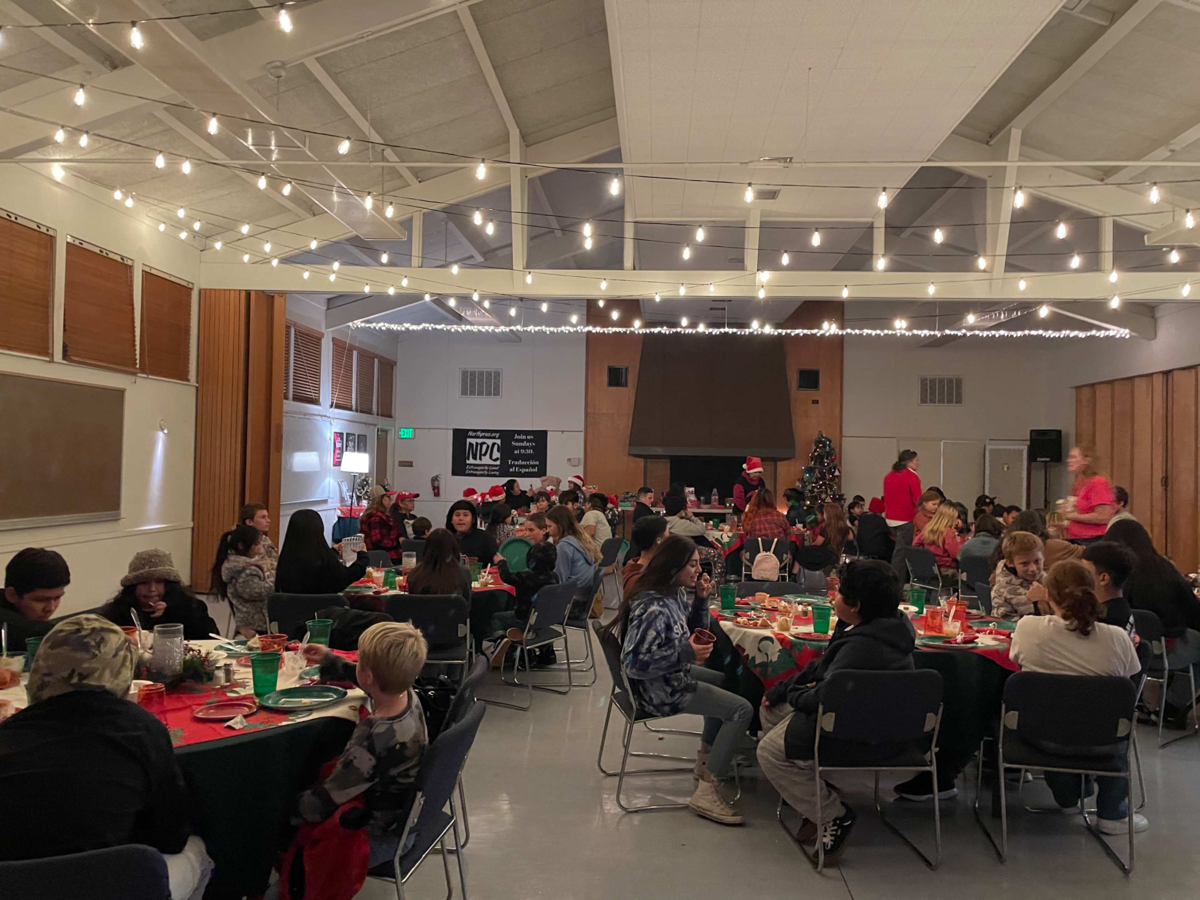 A large indoor space is filled with tables of people gathering for a holiday event, decorated with string lights and a Christmas tree.