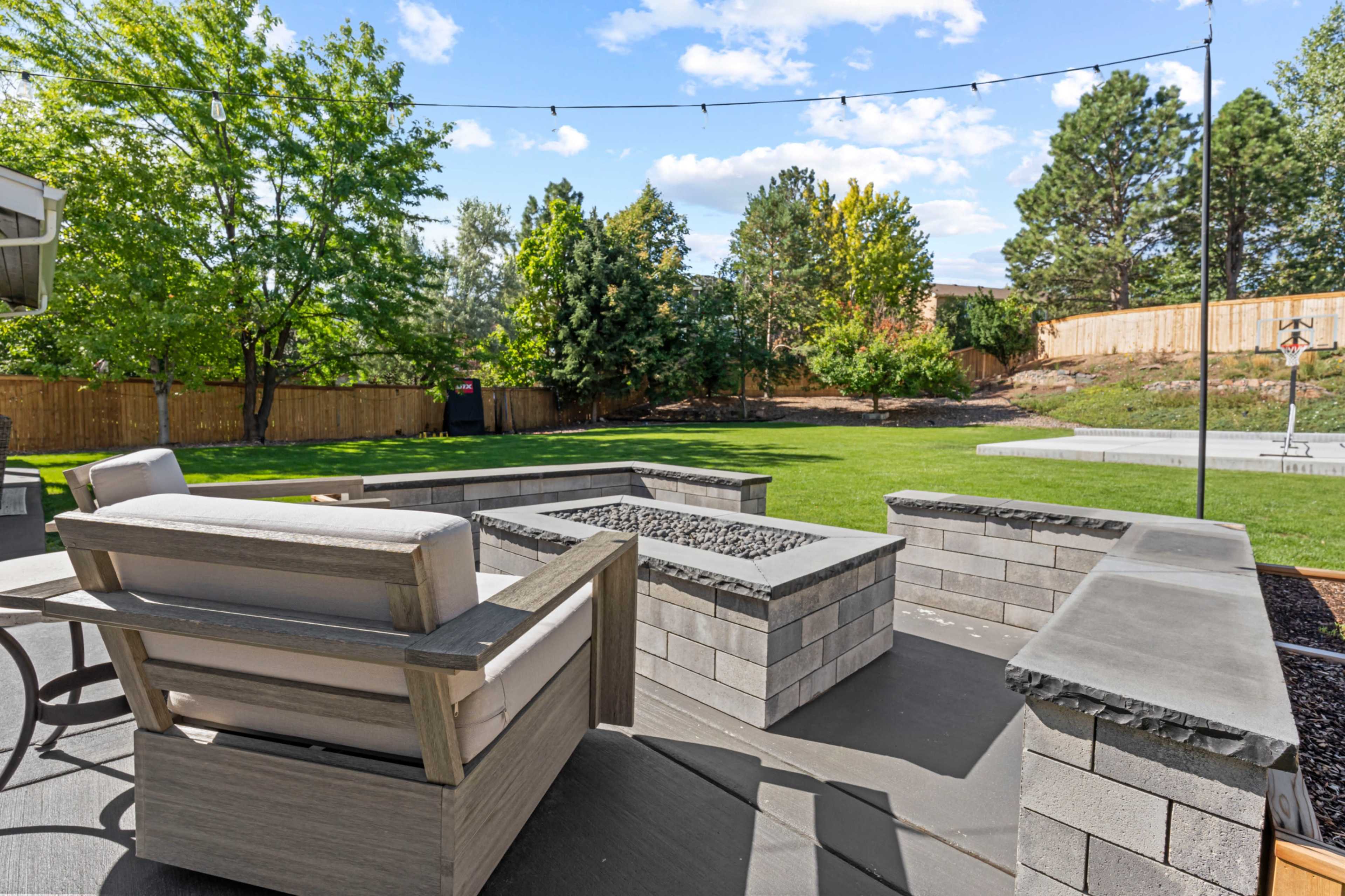 The scene shows a backyard with a modern seating area, a fire pit made of stone, and a well-maintained lawn surrounded by trees and a wooden fence.
