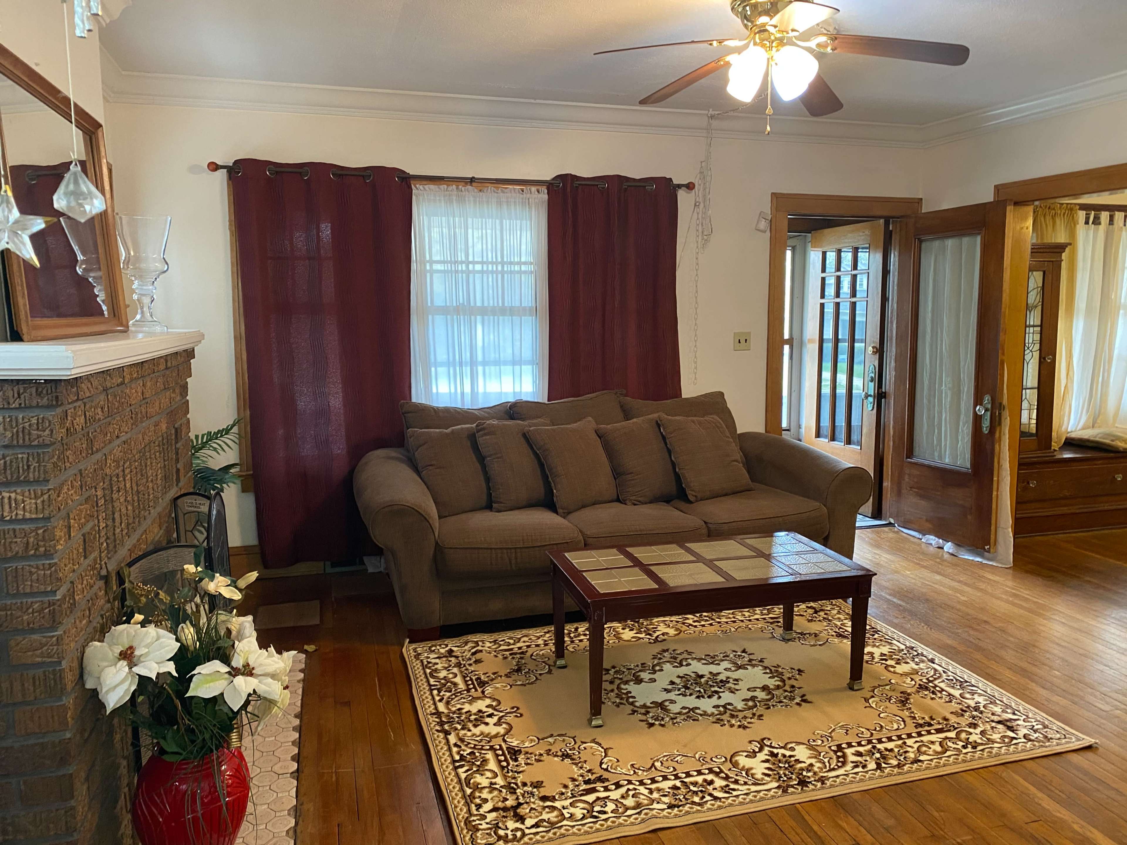 A living room features a brown couch, a glass-top coffee table, and a decorative area rug, with curtains framing a window and a ceiling fan above.