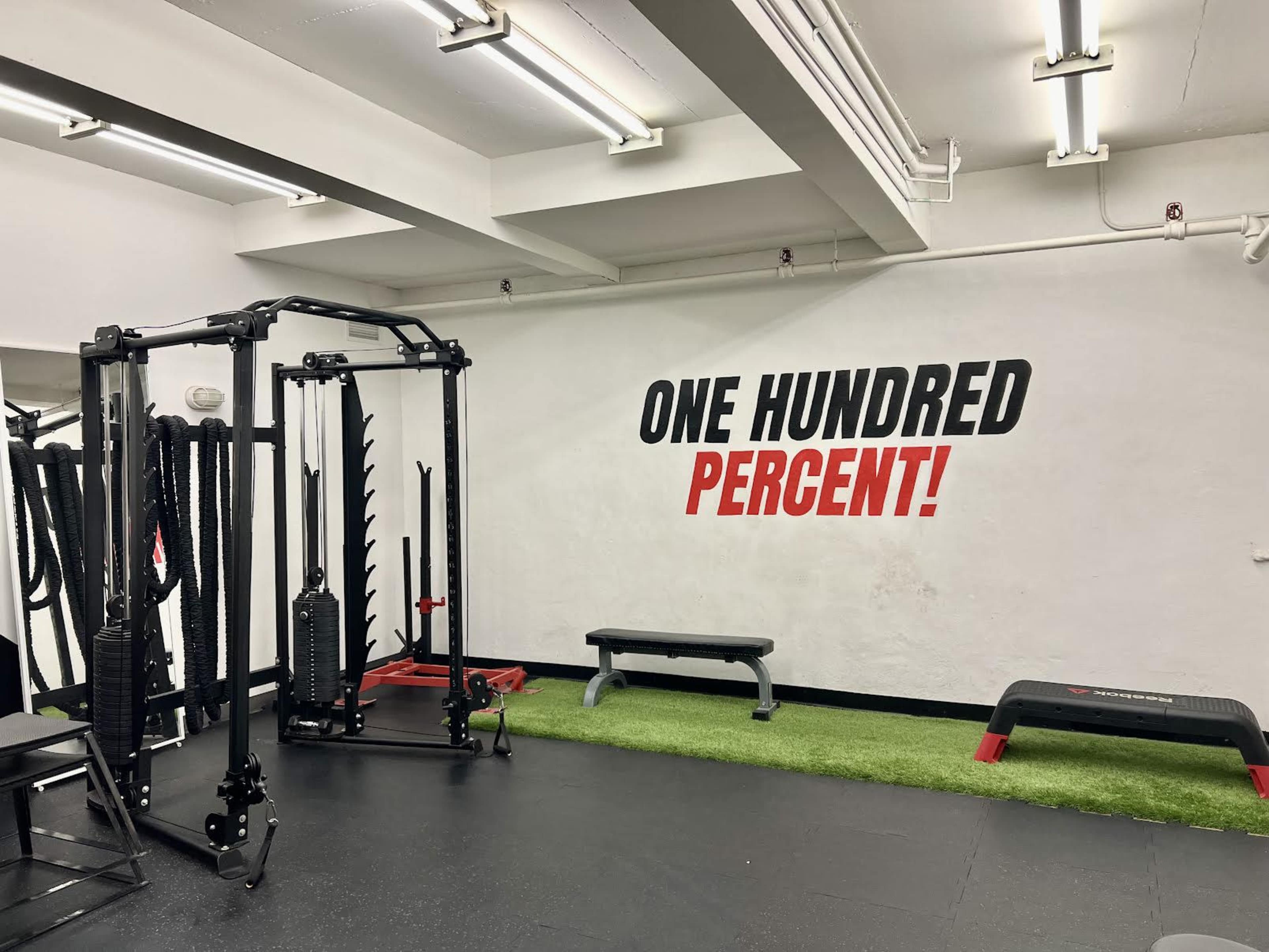 A gym interior with exercise equipment, a bench, and a wall displaying the phrase "ONE HUNDRED PERCENT!" in bold letters.