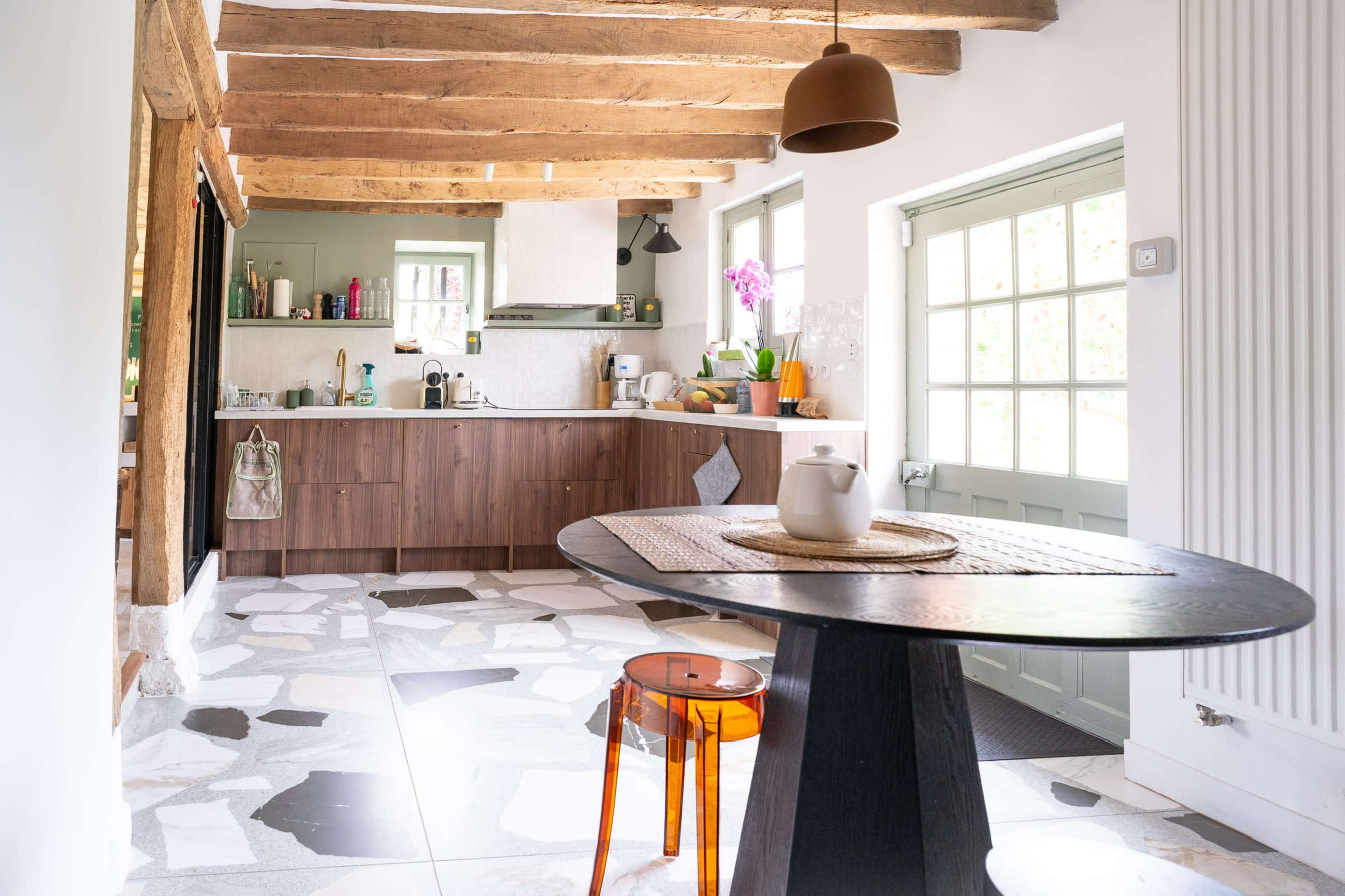 A kitchen area features wooden beams, a two-tone cabinet, and a round table with a transparent orange stool.