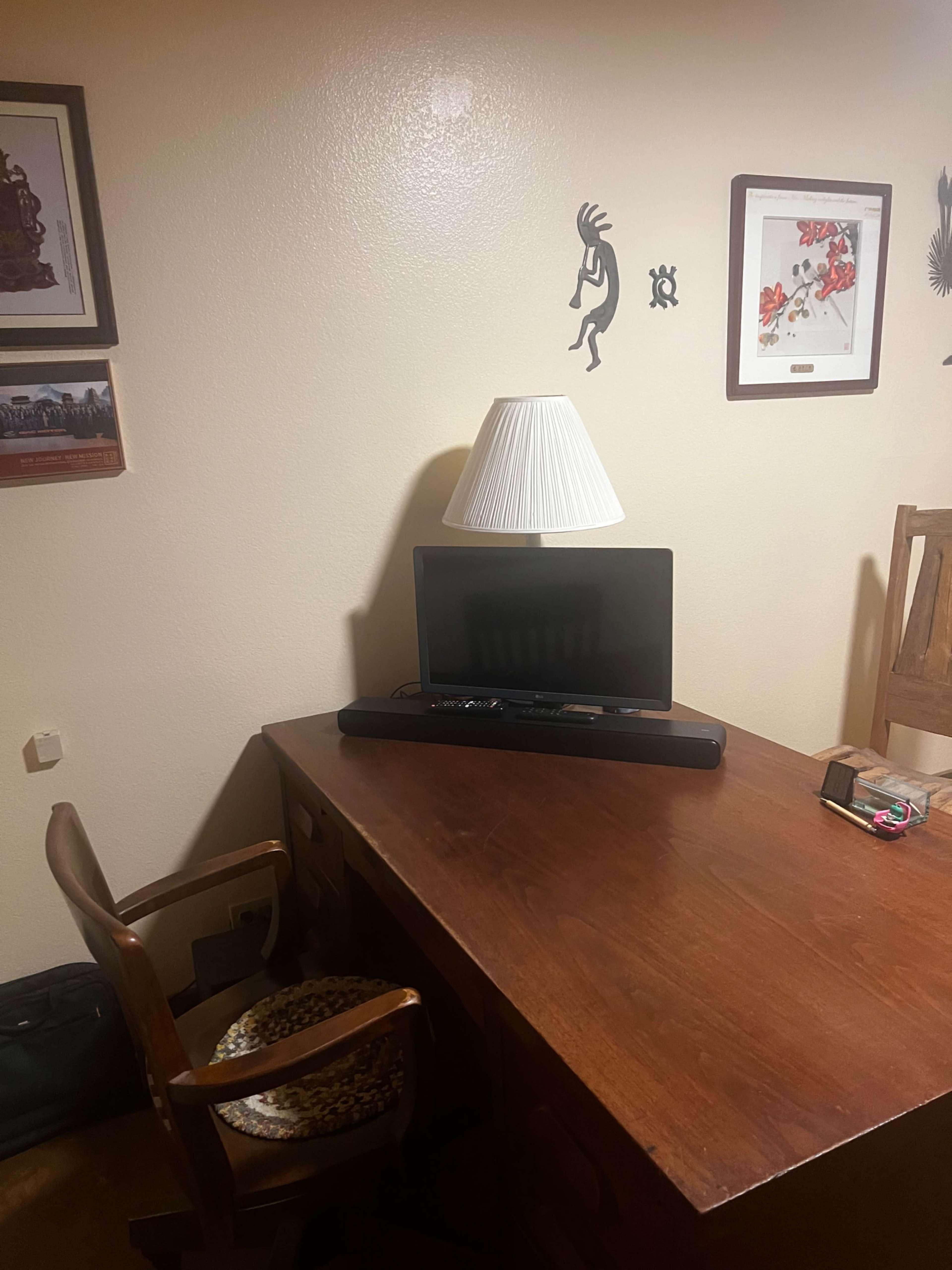 The image shows a wooden desk with a chair, a lamp, a television, and framed artwork on the walls.