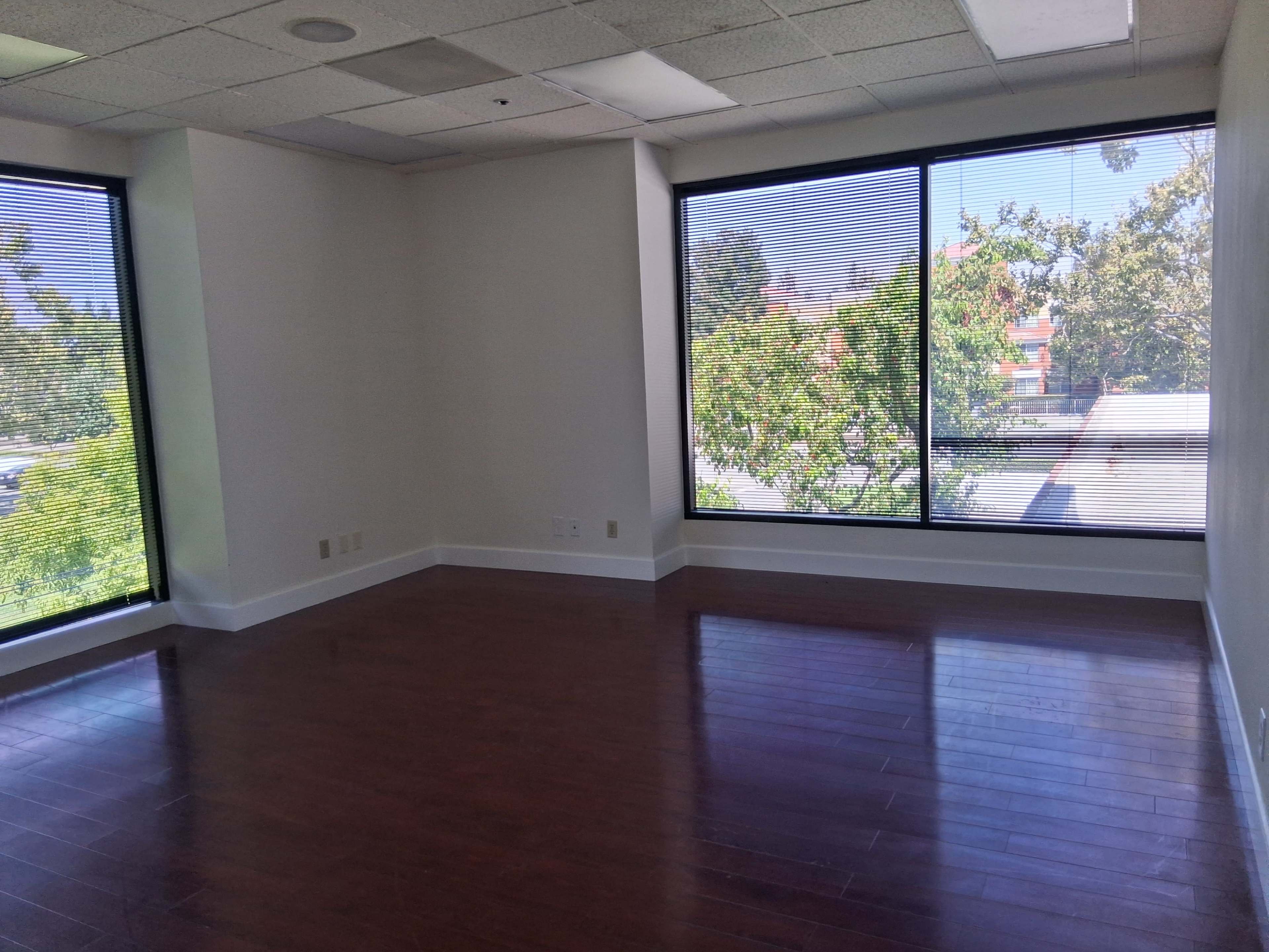 The image shows an empty office space with large windows providing natural light and a view of trees outside.