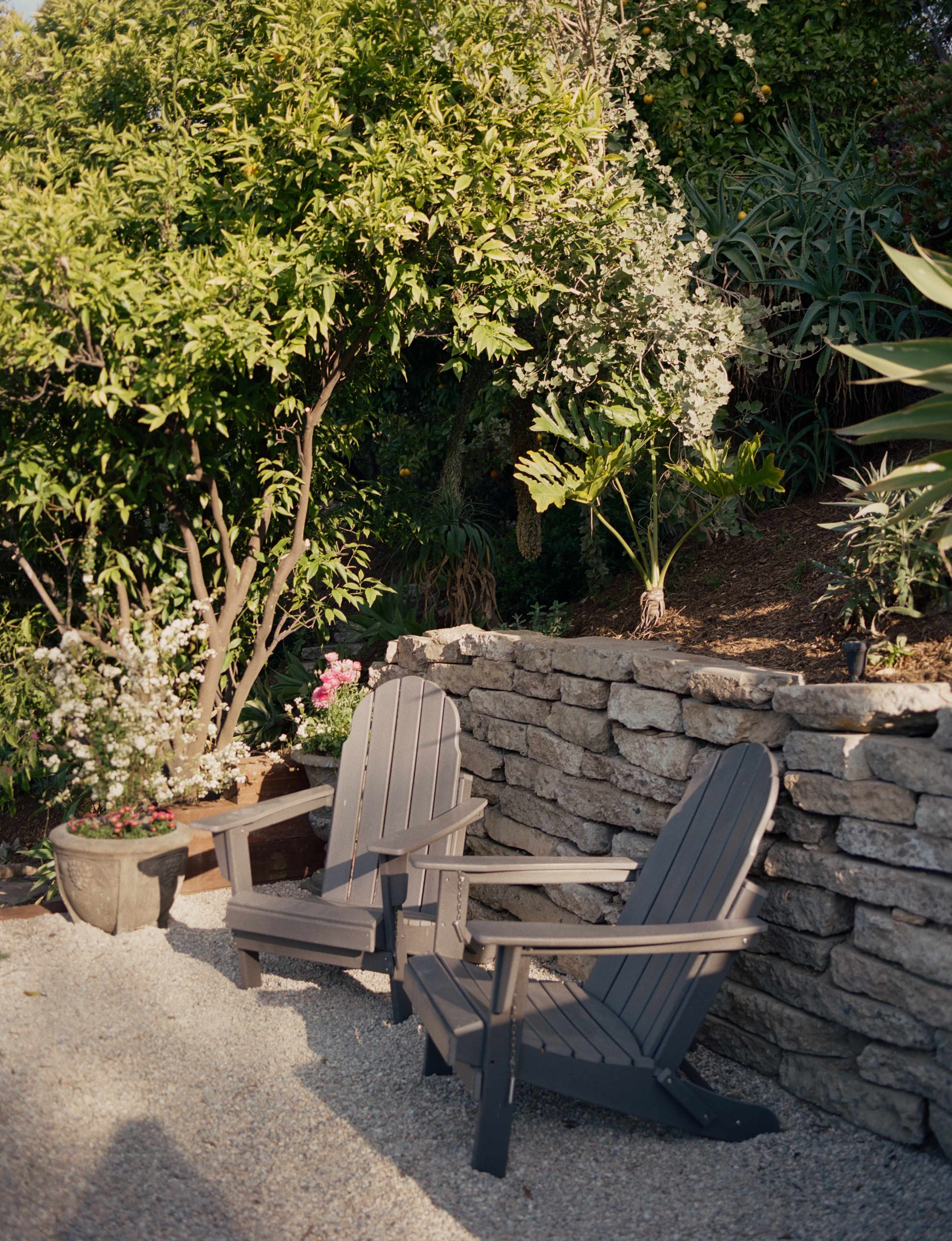 Two wooden Adirondack chairs sit beside a stone wall adorned with various plants and flowers in a garden setting.