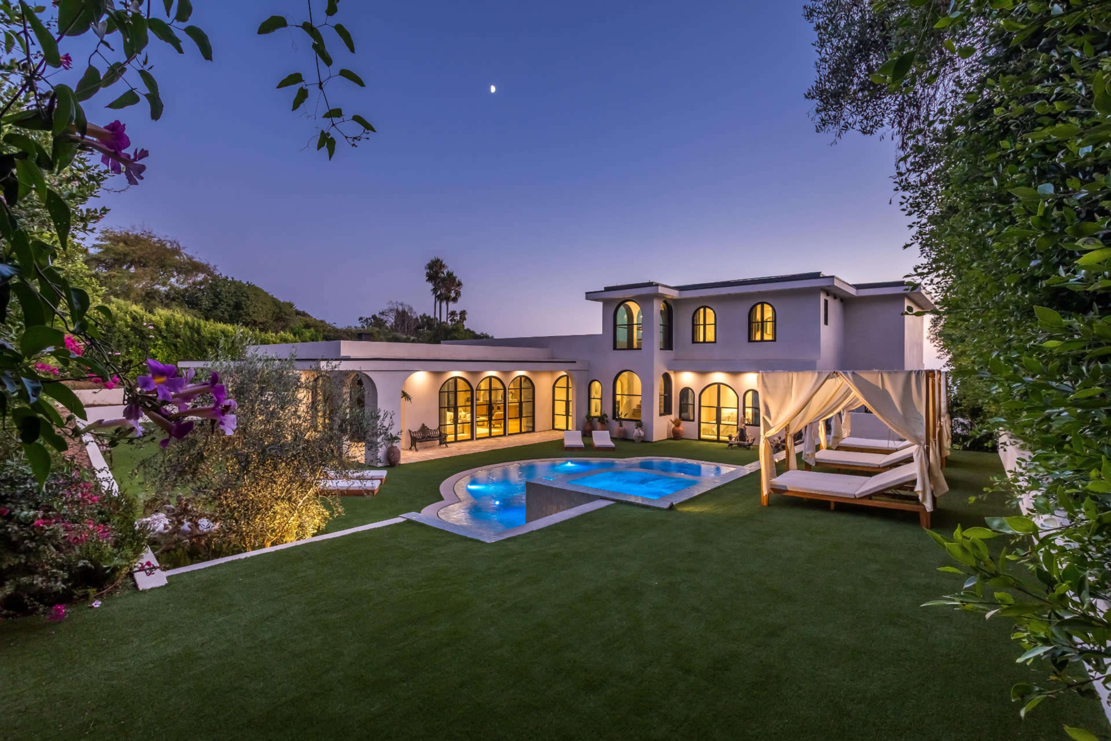 The image shows a modern villa with arched windows, a swimming pool, and landscaped gardens, illuminated in the evening light.