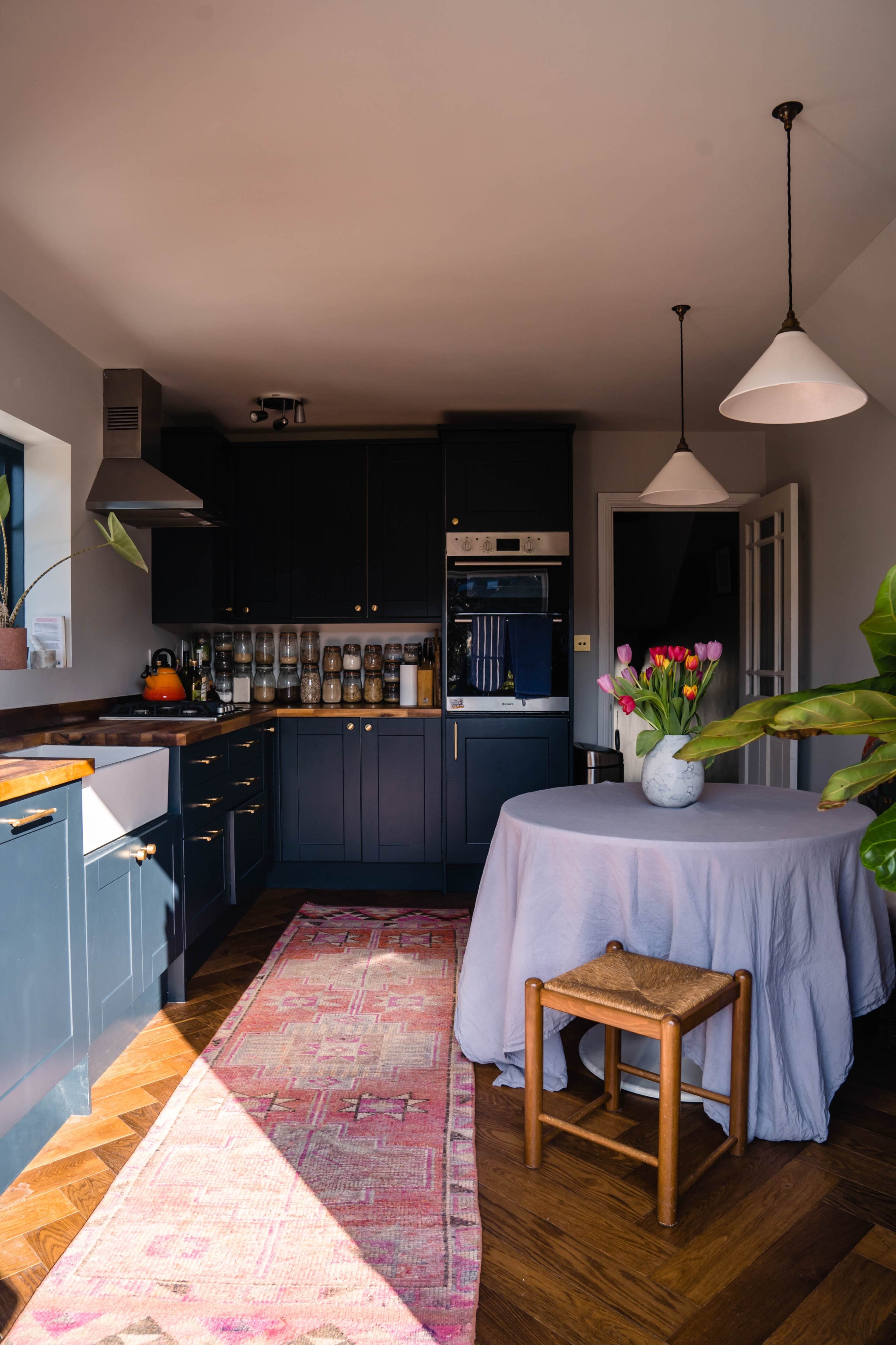 A modern kitchen features dark cabinetry, a round table with a tablecloth and a vase of tulips, and a patterned rug on the hardwood floor.