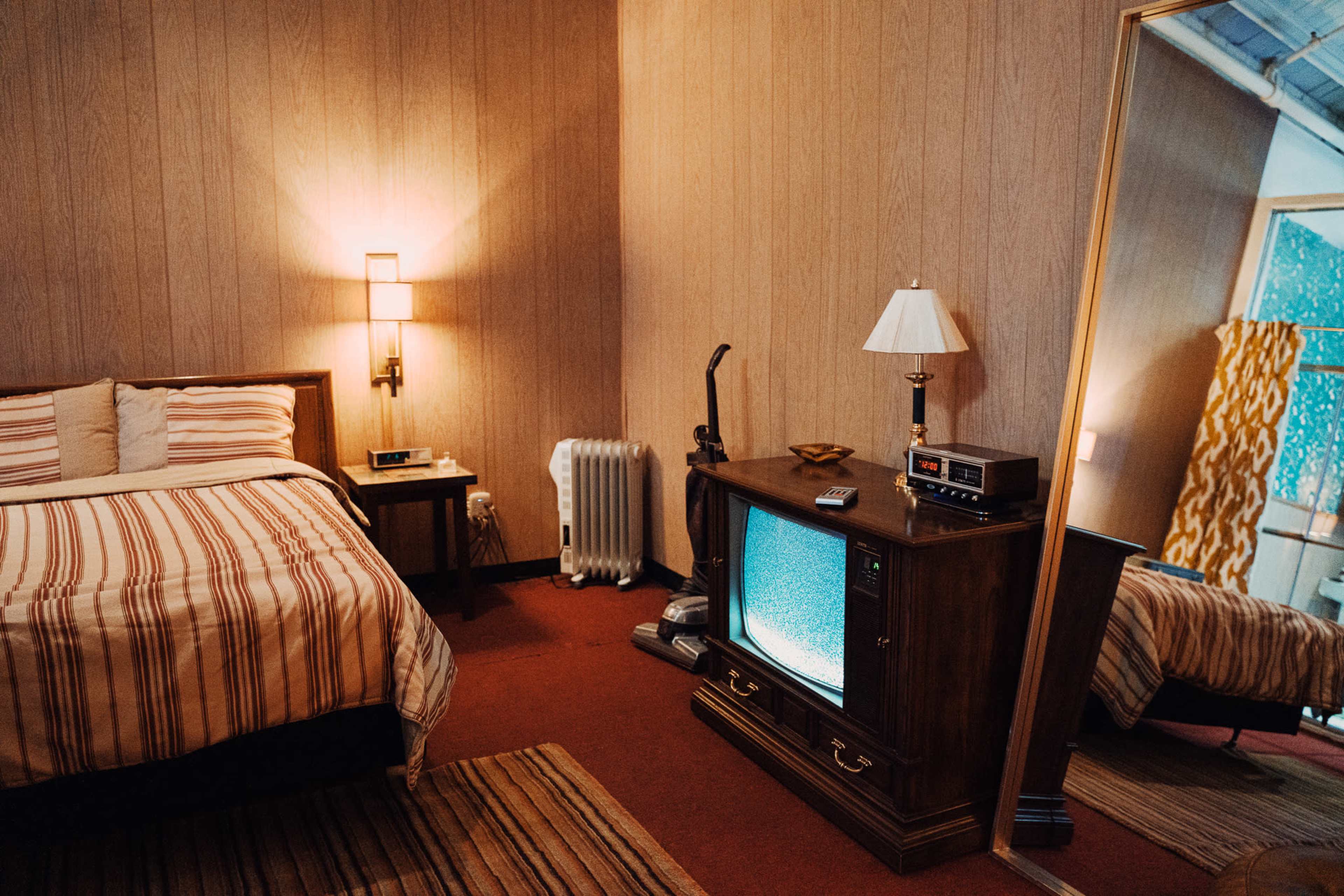 A neatly arranged motel room features a striped bedspread, a vintage television on a wooden stand, a lamp, and a mirror reflecting part of the room.
