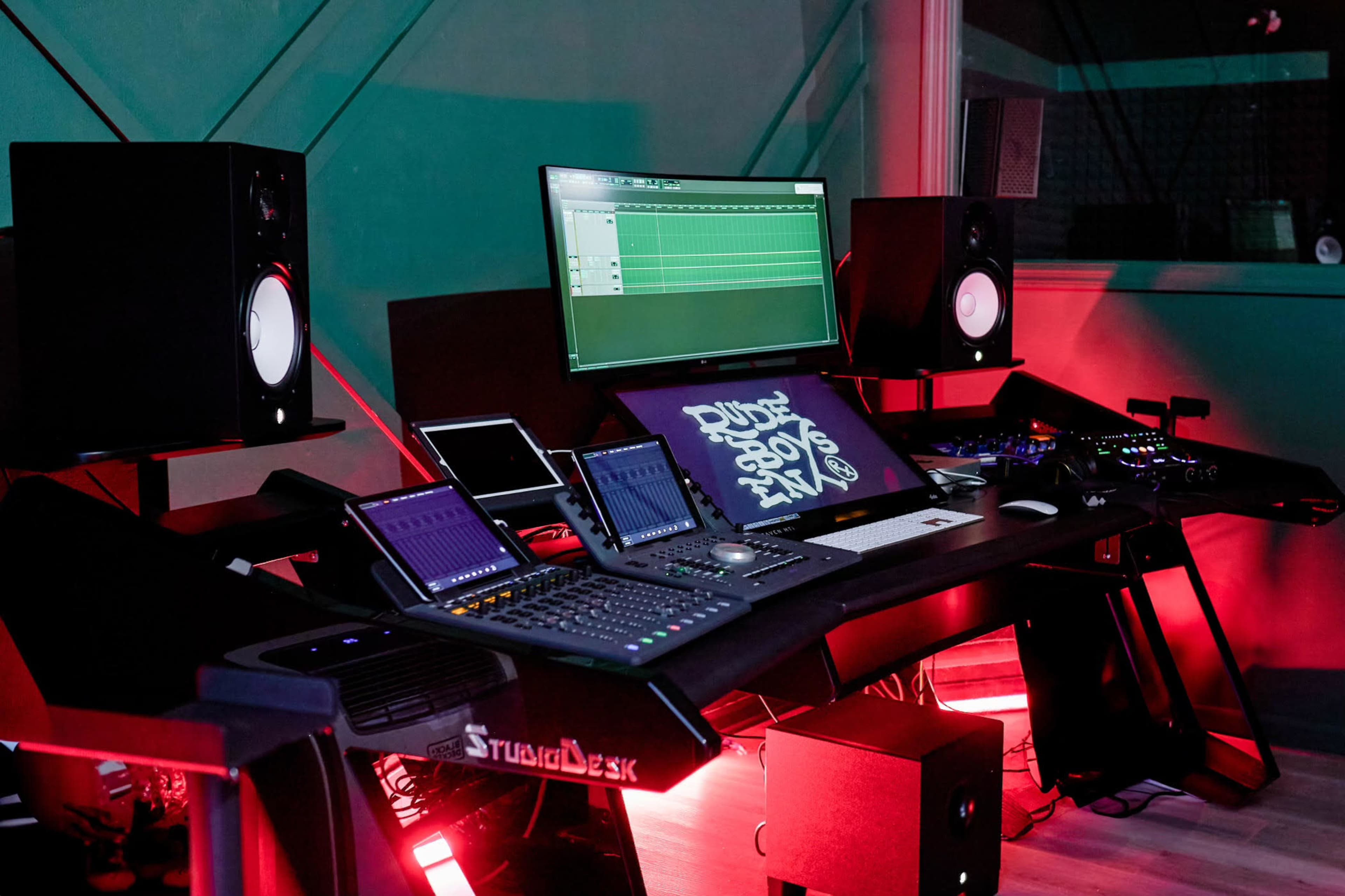 The image shows a modern music production studio with a central workstation, multiple screens, audio speakers, and colorful lighting.