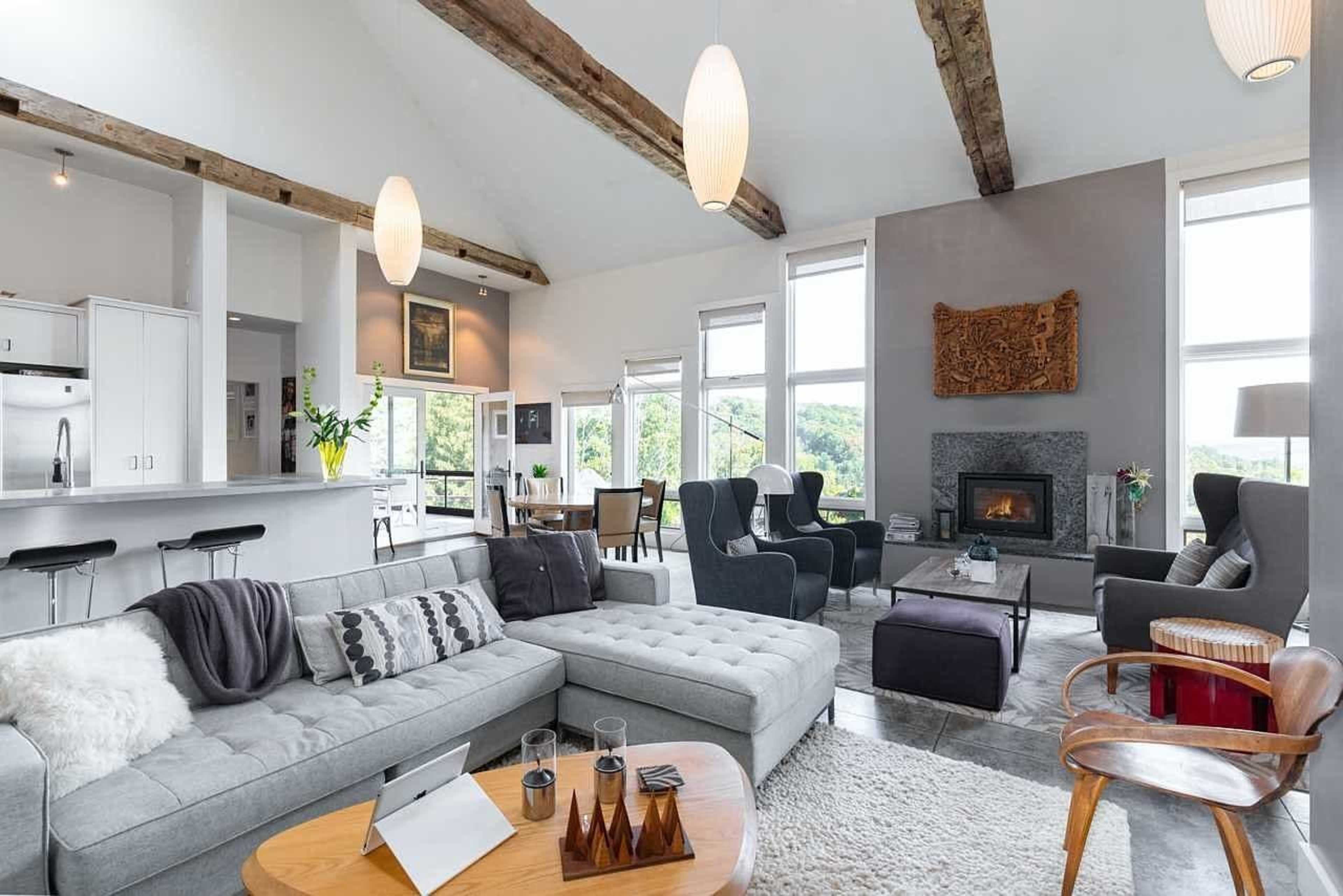 A spacious living area features a gray sectional sofa, wooden beams, and large windows with a view of the outdoors.