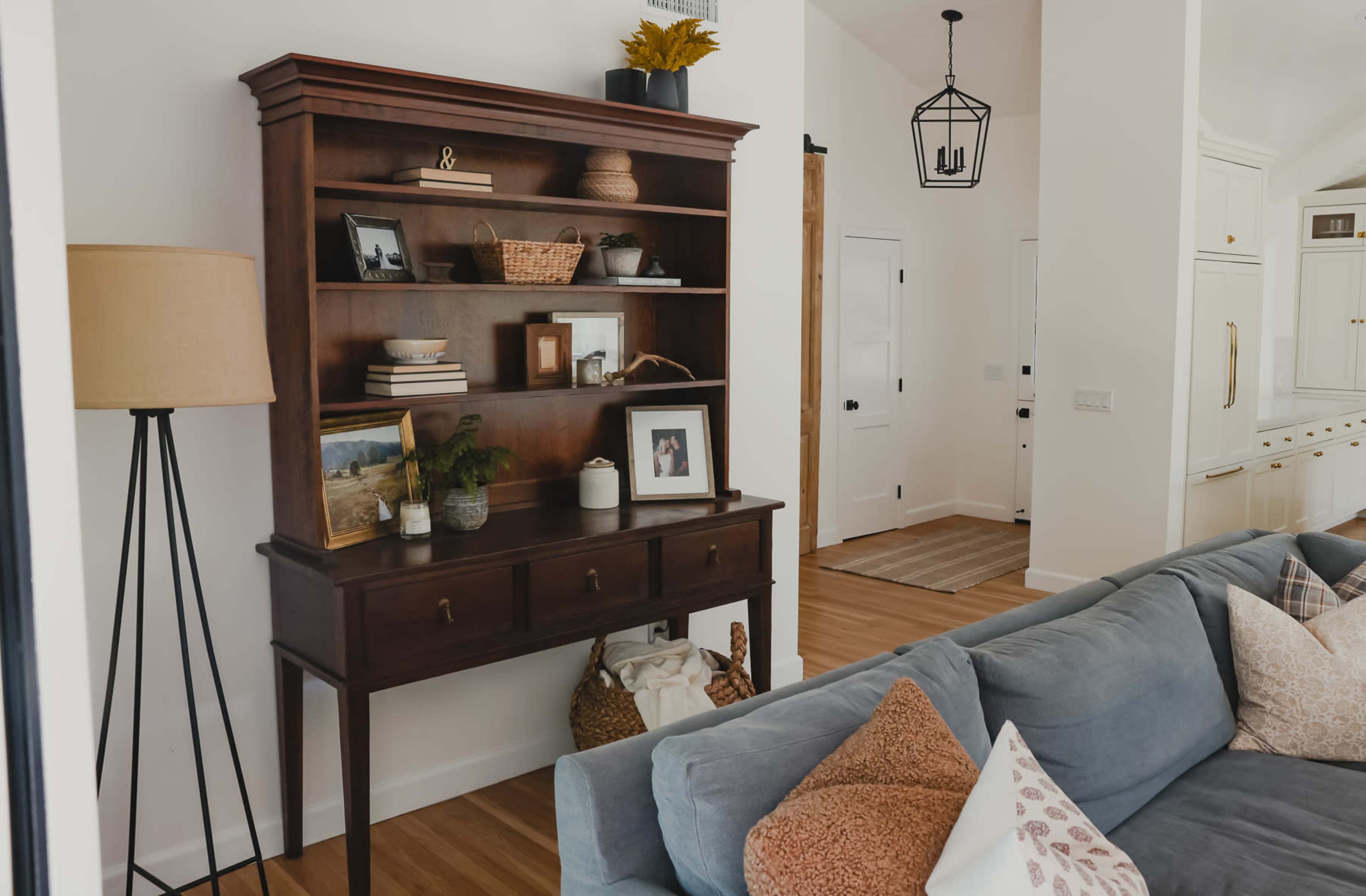 A dark wooden bookshelf with decor and framed photos stands beside a blue couch in a well-lit room with white walls and a simple pendant light.