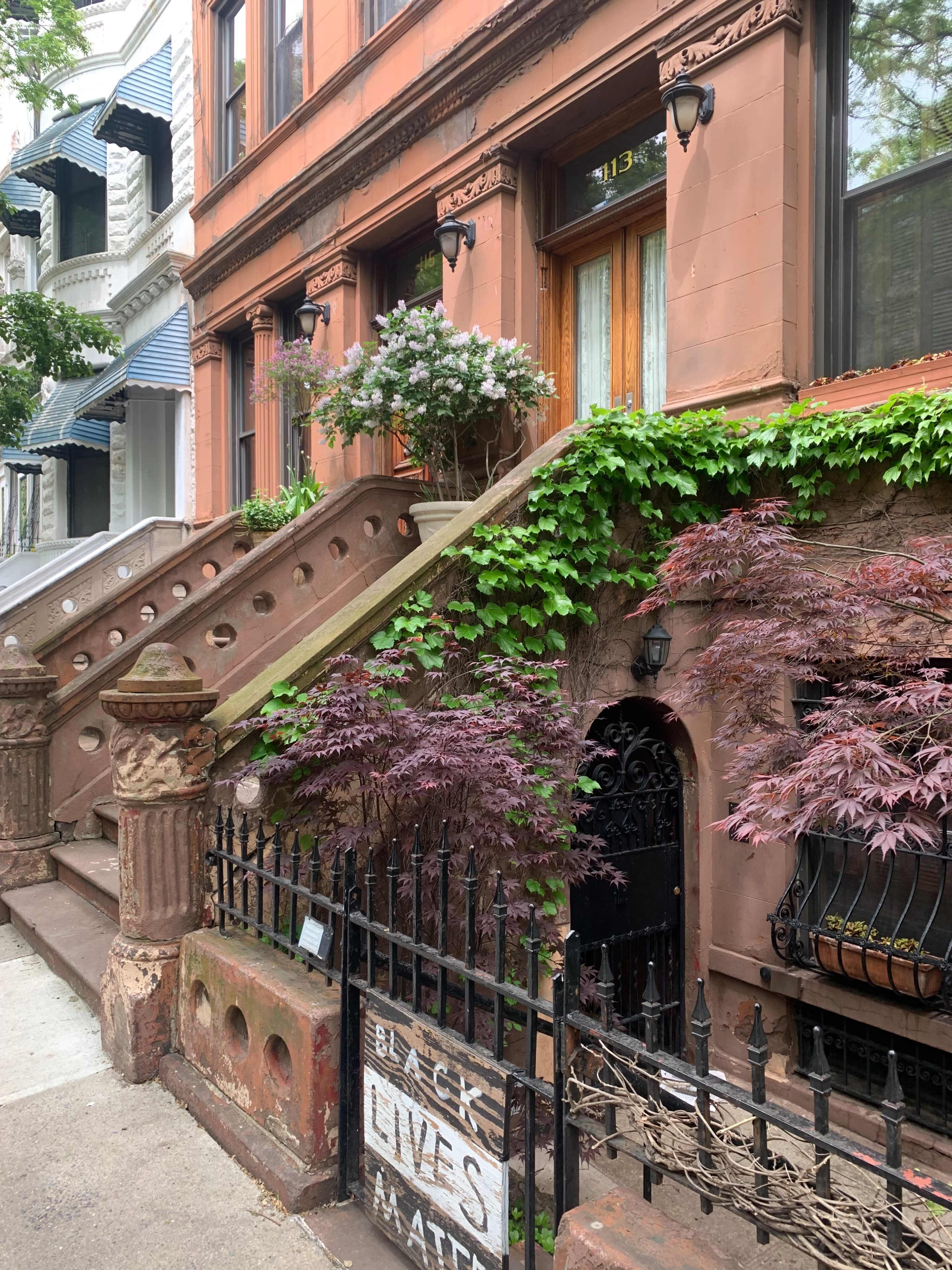 A brownstone building features a wrought iron fence, a prominent "Black Lives Matter" sign, and flowering plants along the steps.