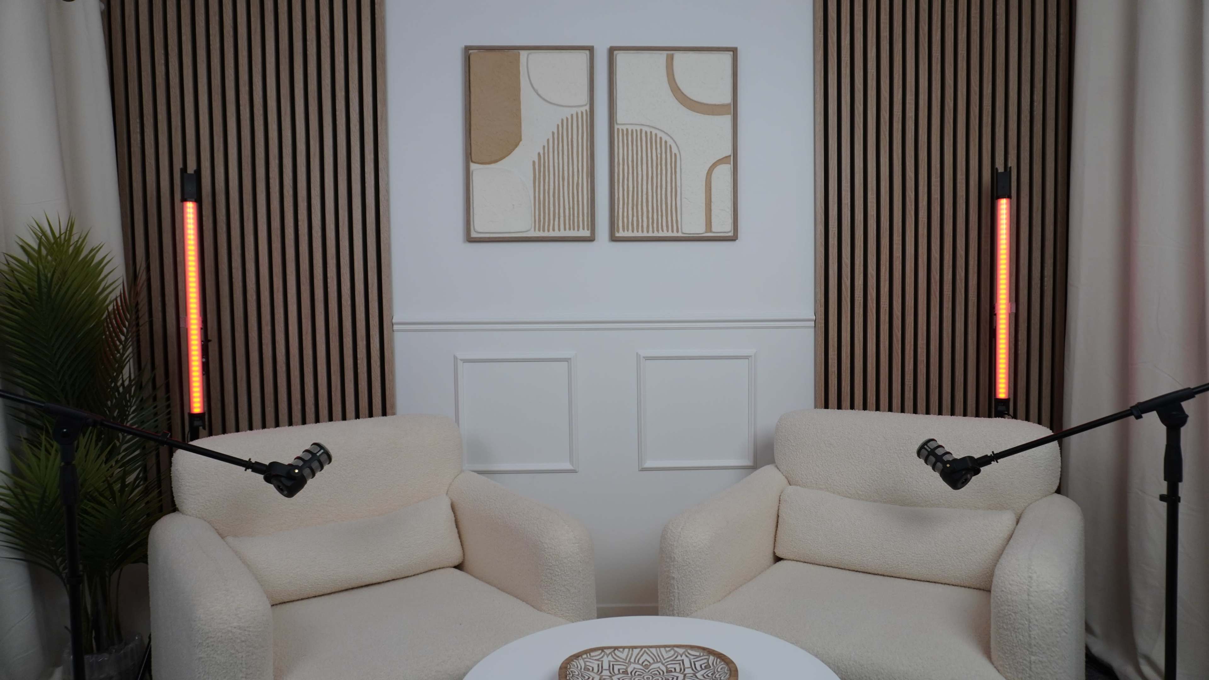 The image shows a minimalist interior with two beige chairs facing each other, a round table in the center, and two microphones mounted on stands beside the chairs, with abstract artwork on the wall.