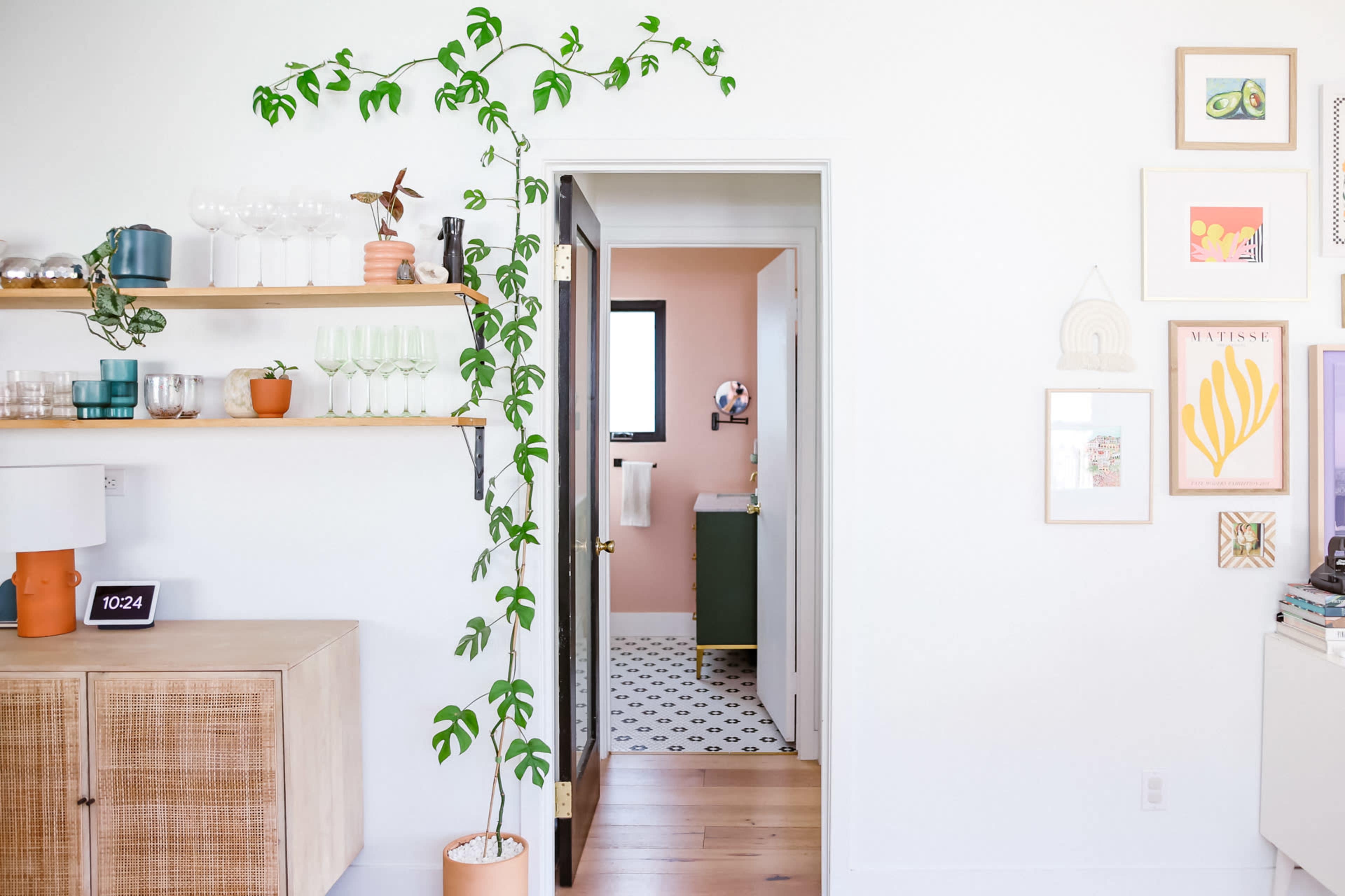 A bright and modern room features a wall-mounted shelf with glassware and plants, leading to a bathroom through an open door.