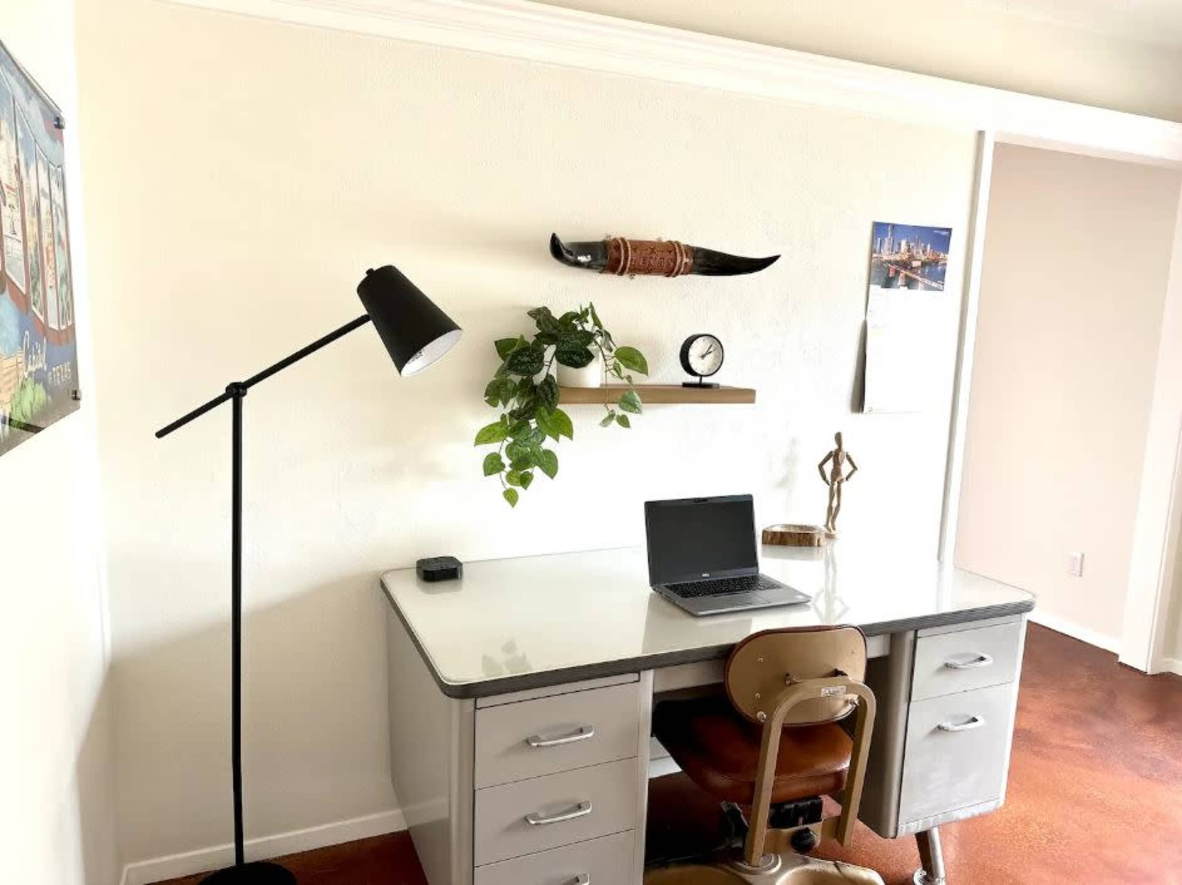 A minimalist workspace features a desk with a laptop, a plant on a shelf, a decorative item, and a standing lamp against a light-colored wall.
