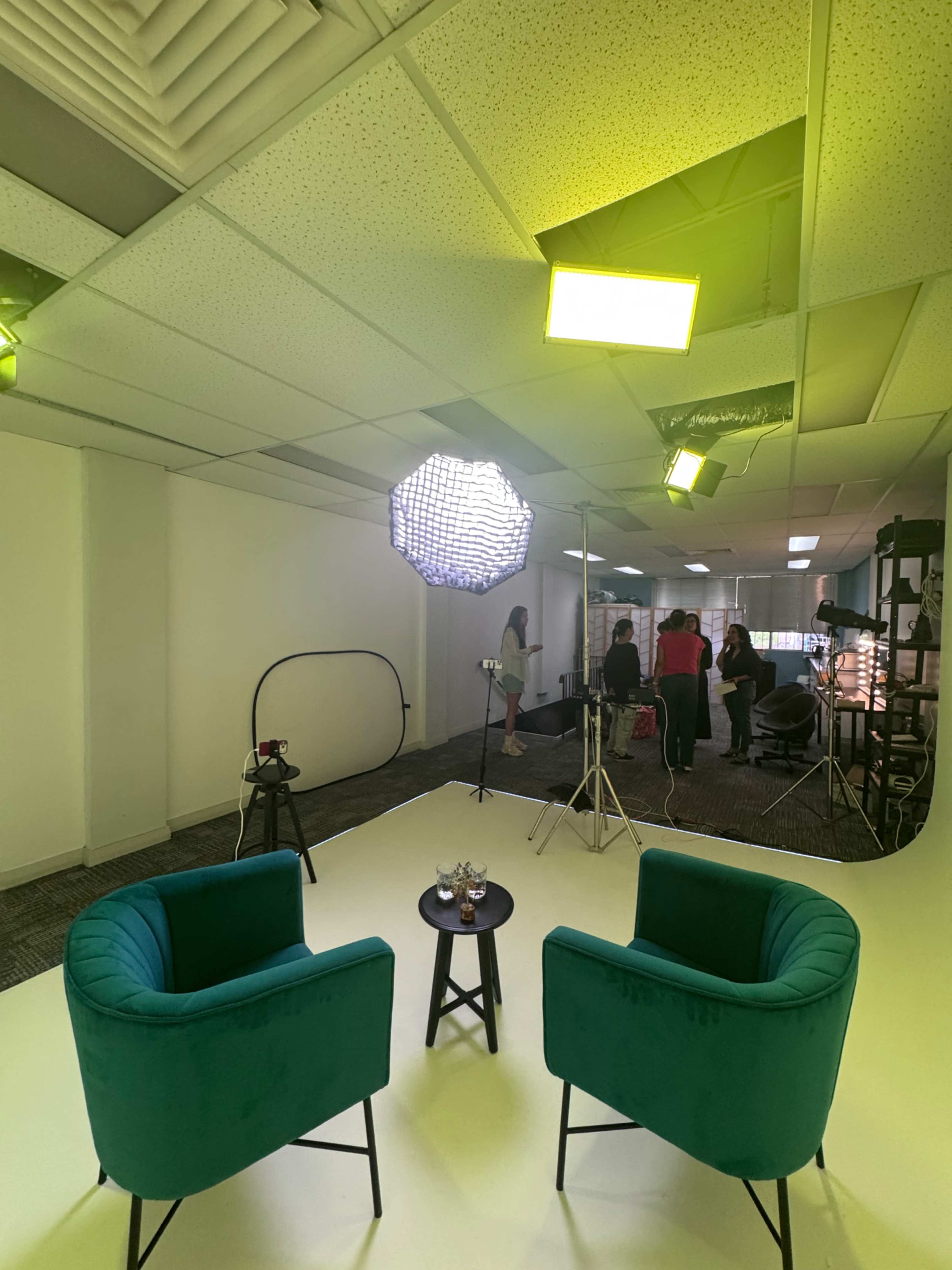 The image shows a studio setup with two green chairs, a small table in the center, and multiple lighting equipment, with some people in the background preparing for a shoot.